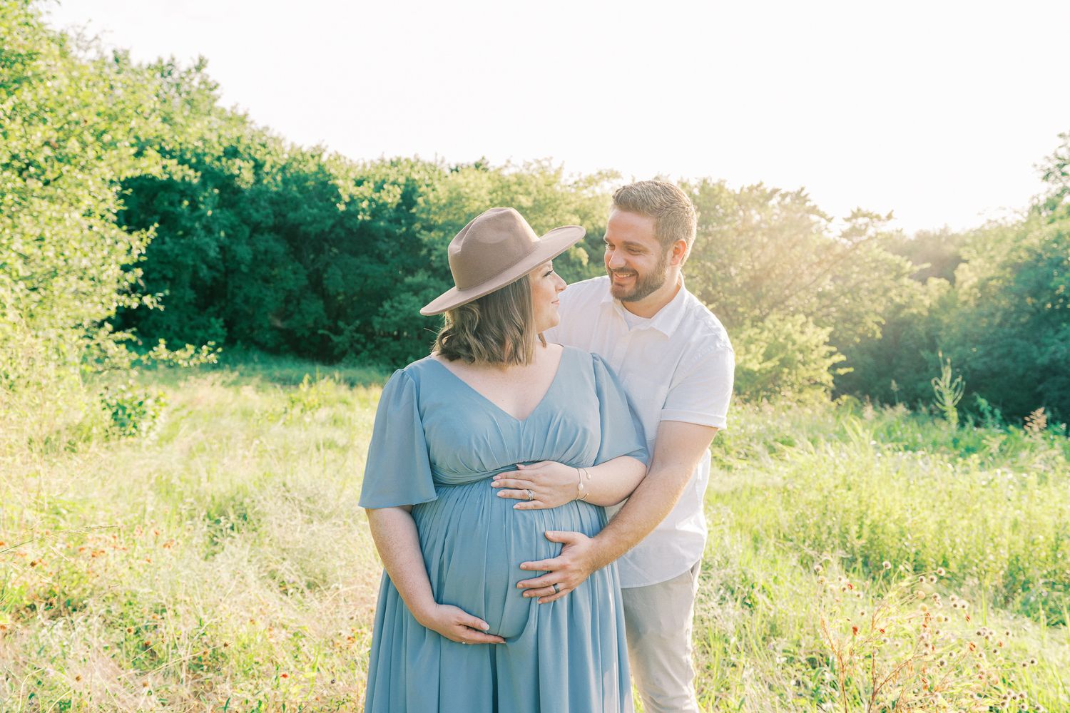 A couple in blue and white outfits embrace in a sunlit meadow during a golden hour photoshoot.