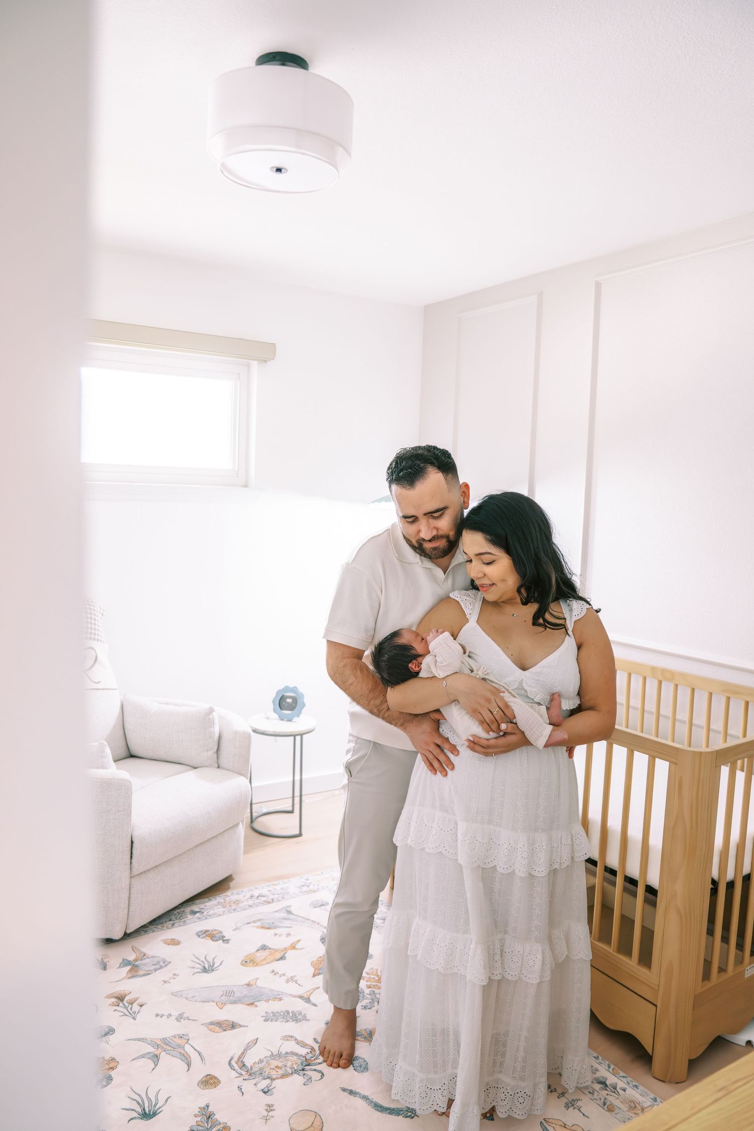 A couple tenderly holds their newborn baby in a bright, white nursery with a wooden crib and floral rug.