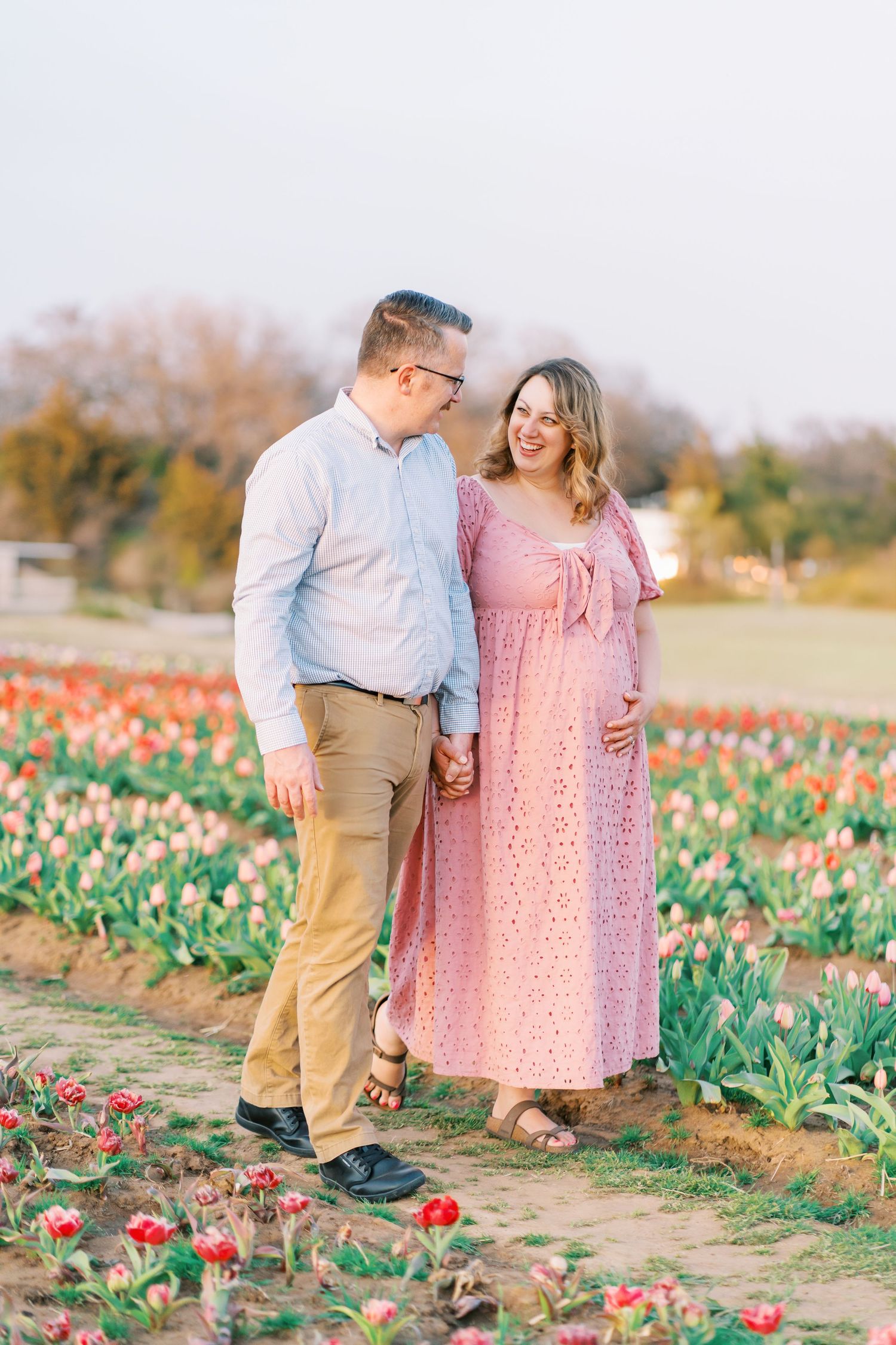 Pregnant couple holding hands in a tulip field, woman in pink dress cradling baby bump, man in blue shirt smiling at her.