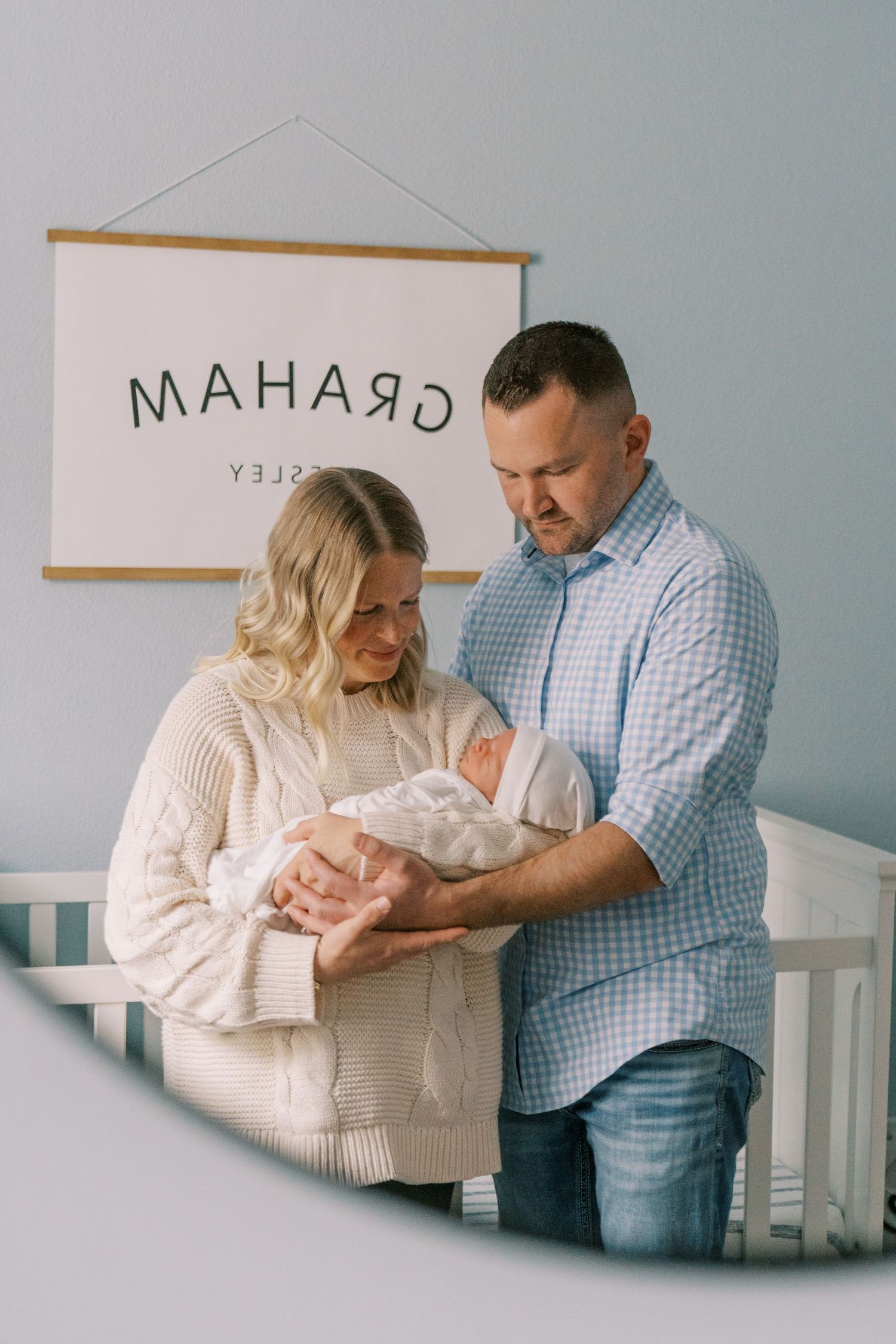 New parents tenderly hold their newborn baby in a nursery with a name sign reading Graham on the wall.