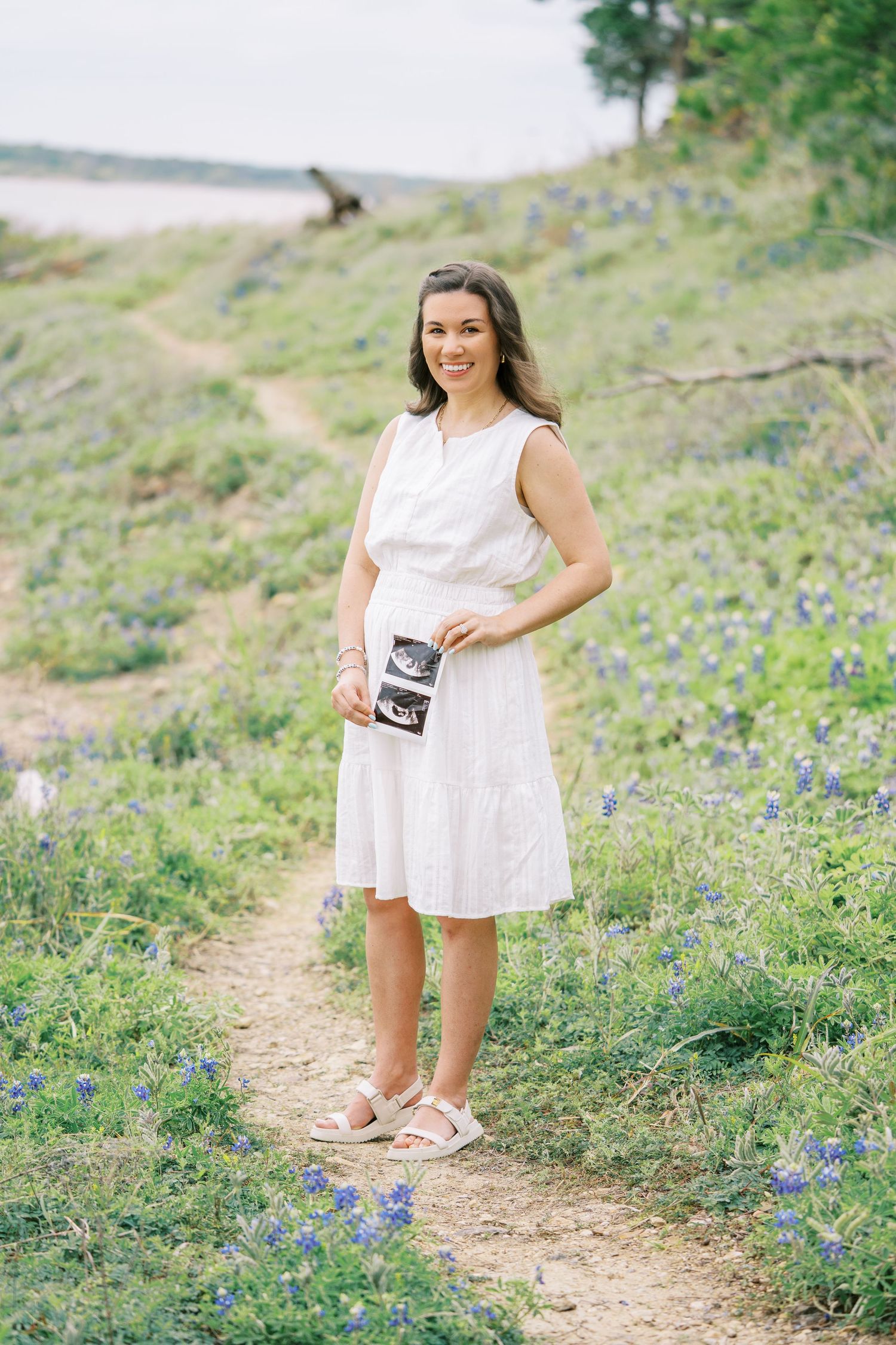 Smiling pregnant woman in white dress holding ultrasound photos on a wildflower trail with blue bonnets in bloom.