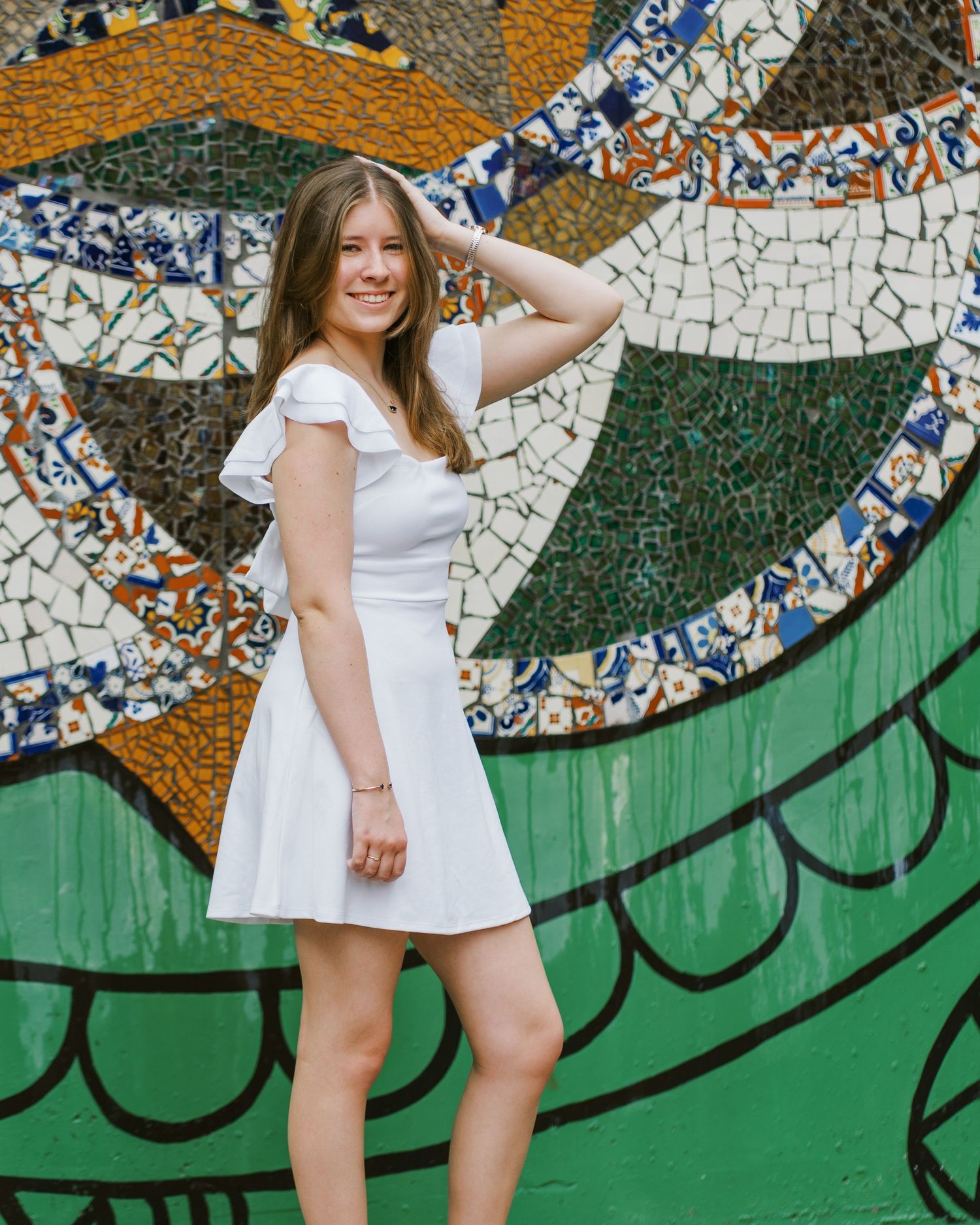 A person in a white dress poses playfully against a mosaic wall with green and orange artistic patterns.