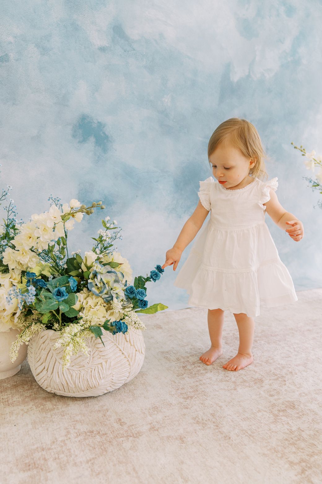 A small child in a white dress explores a delicate floral arrangement on a light backdrop.