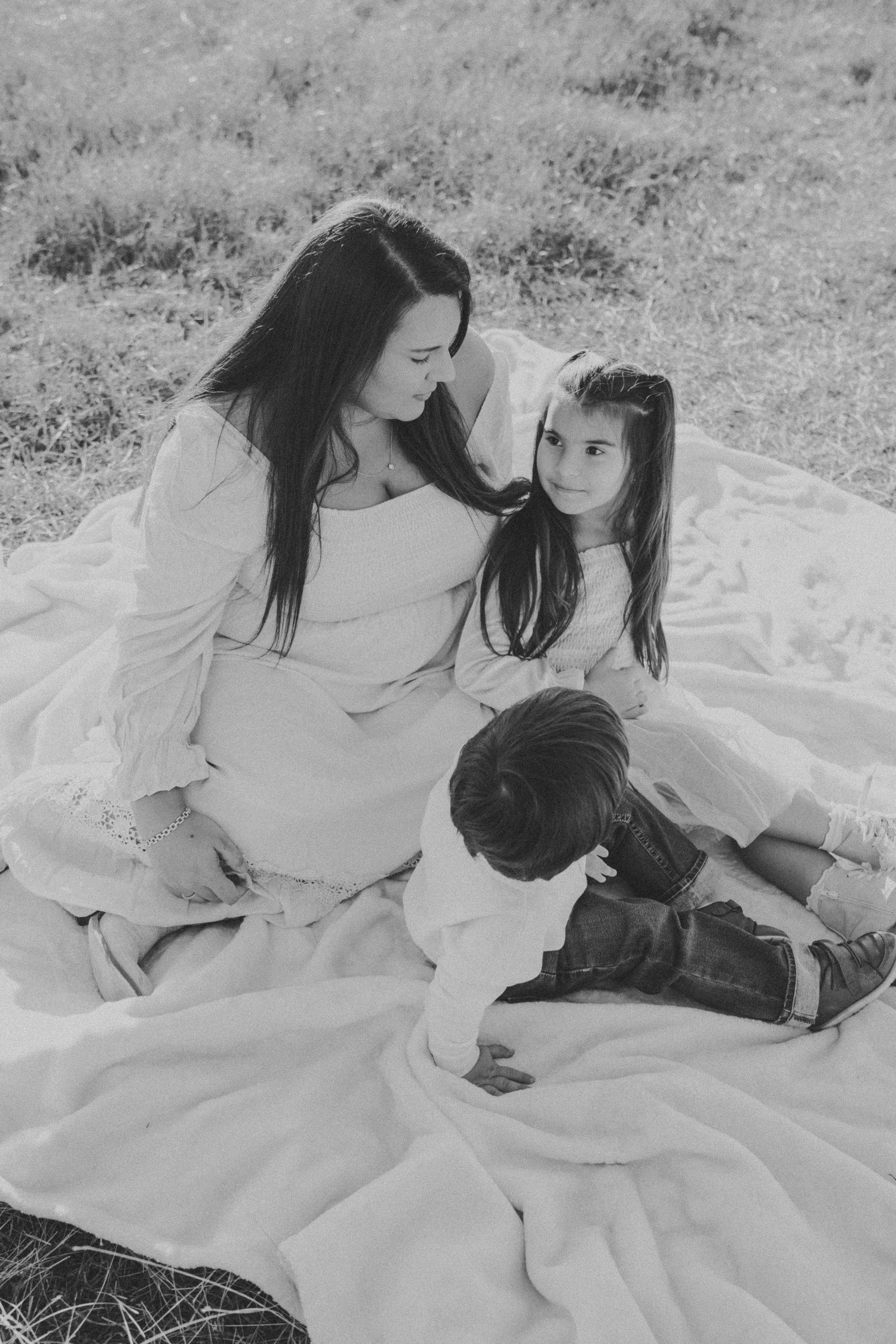 Black and white photo of a tender family moment on a white blanket outdoors.