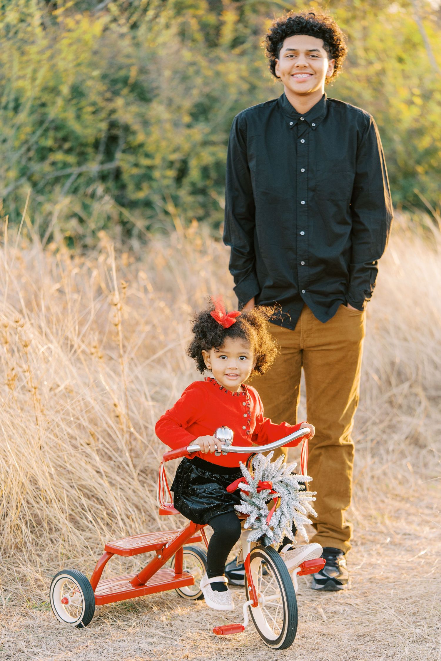 A small child rides a red tricycle on a dirt path while an adult stands nearby in autumn golden light.