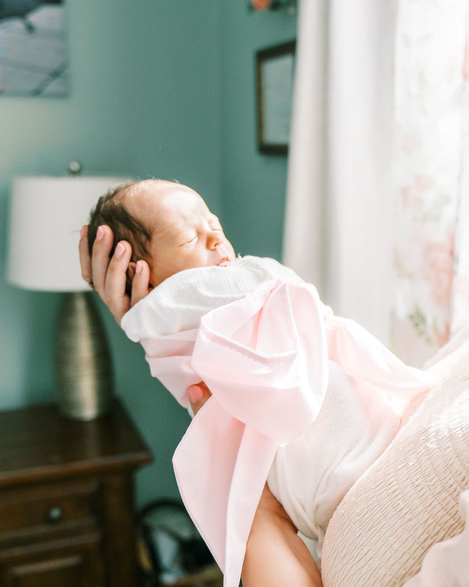 A sleeping newborn wrapped in pink and white being cradled in natural window light.