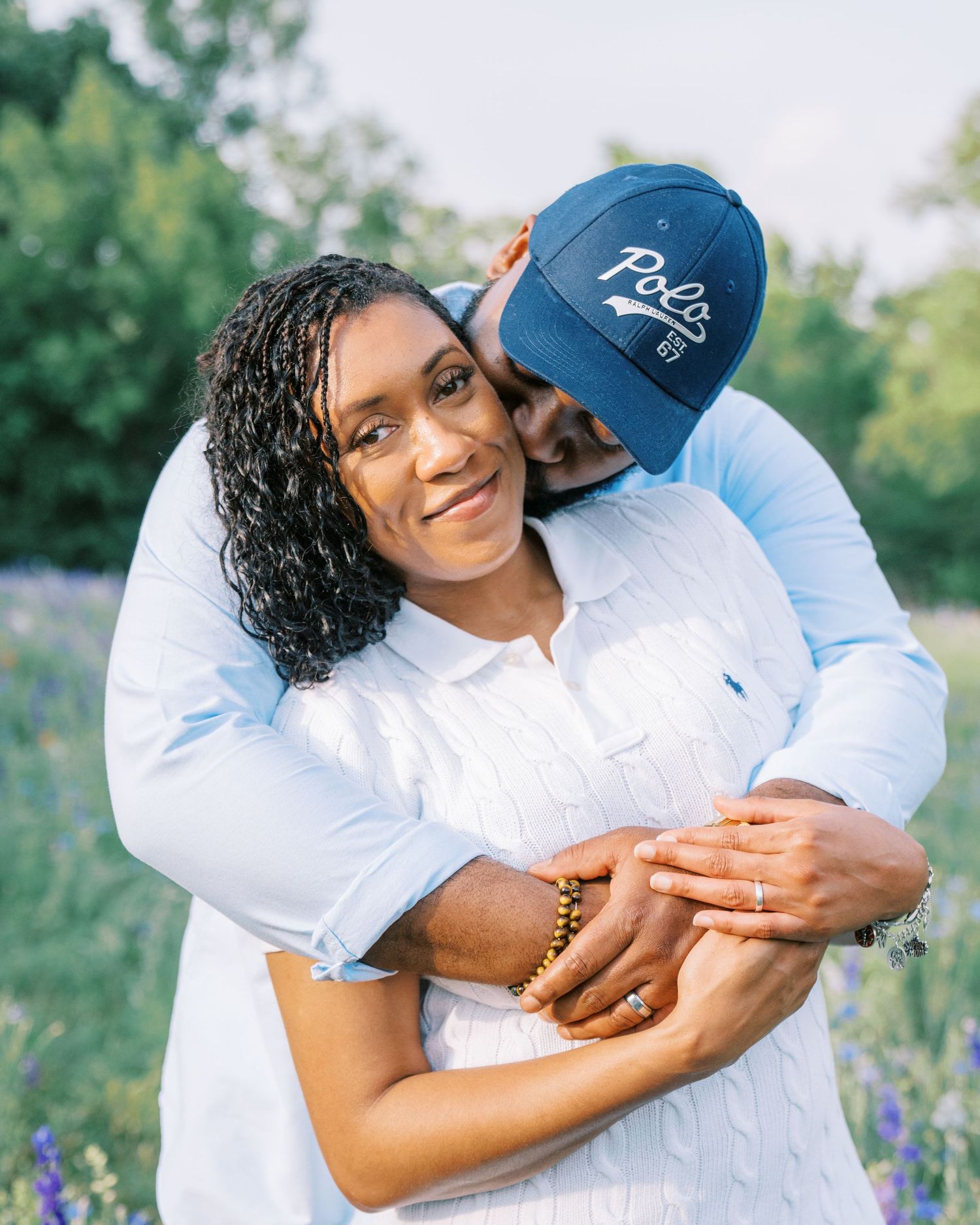 A couple embraces outdoors, woman smiling in white shirt, man in blue Polo cap kissing her cheek in a flower field.