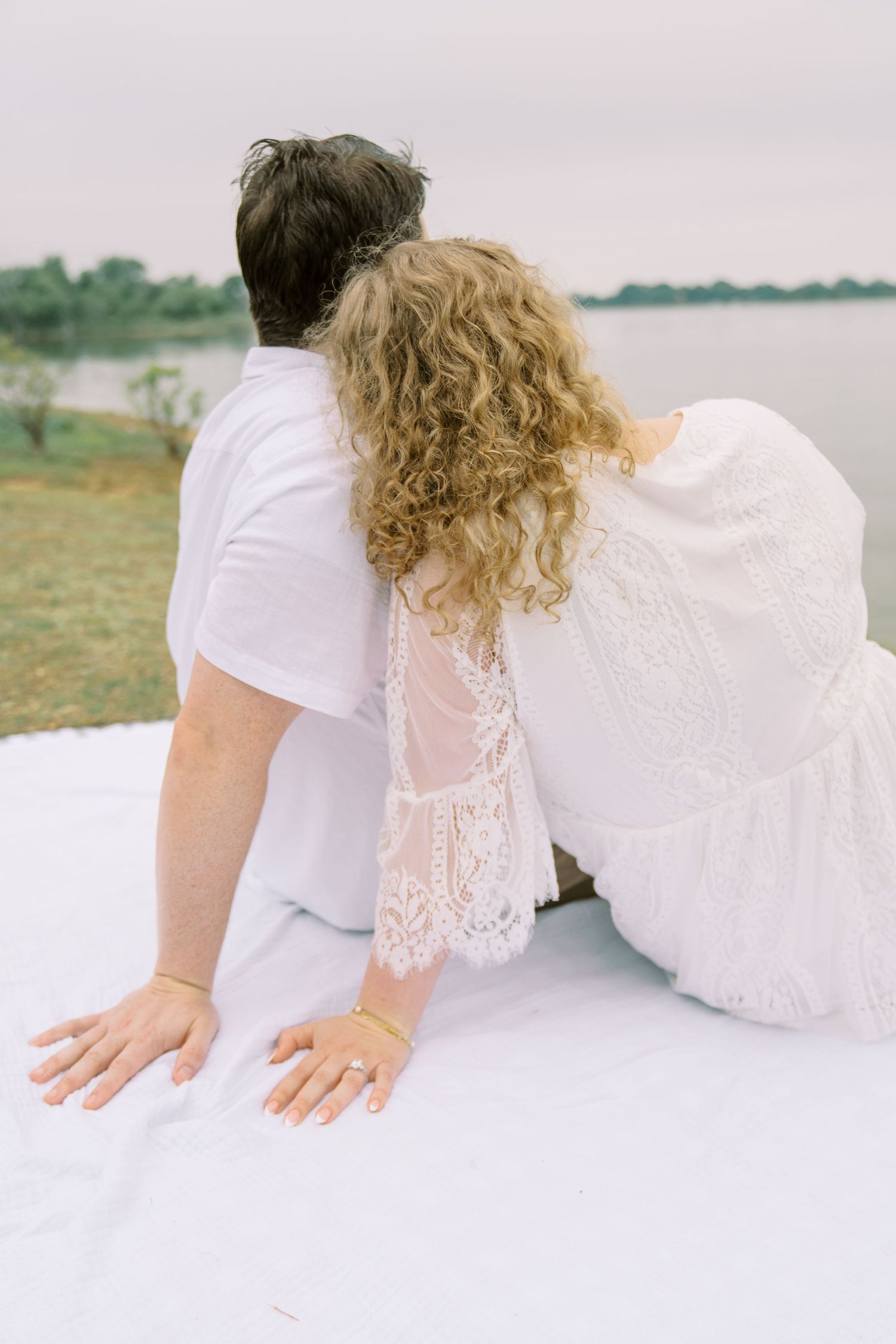 Couple in white sitting on a blanket overlooking a serene lake, viewed from behind.