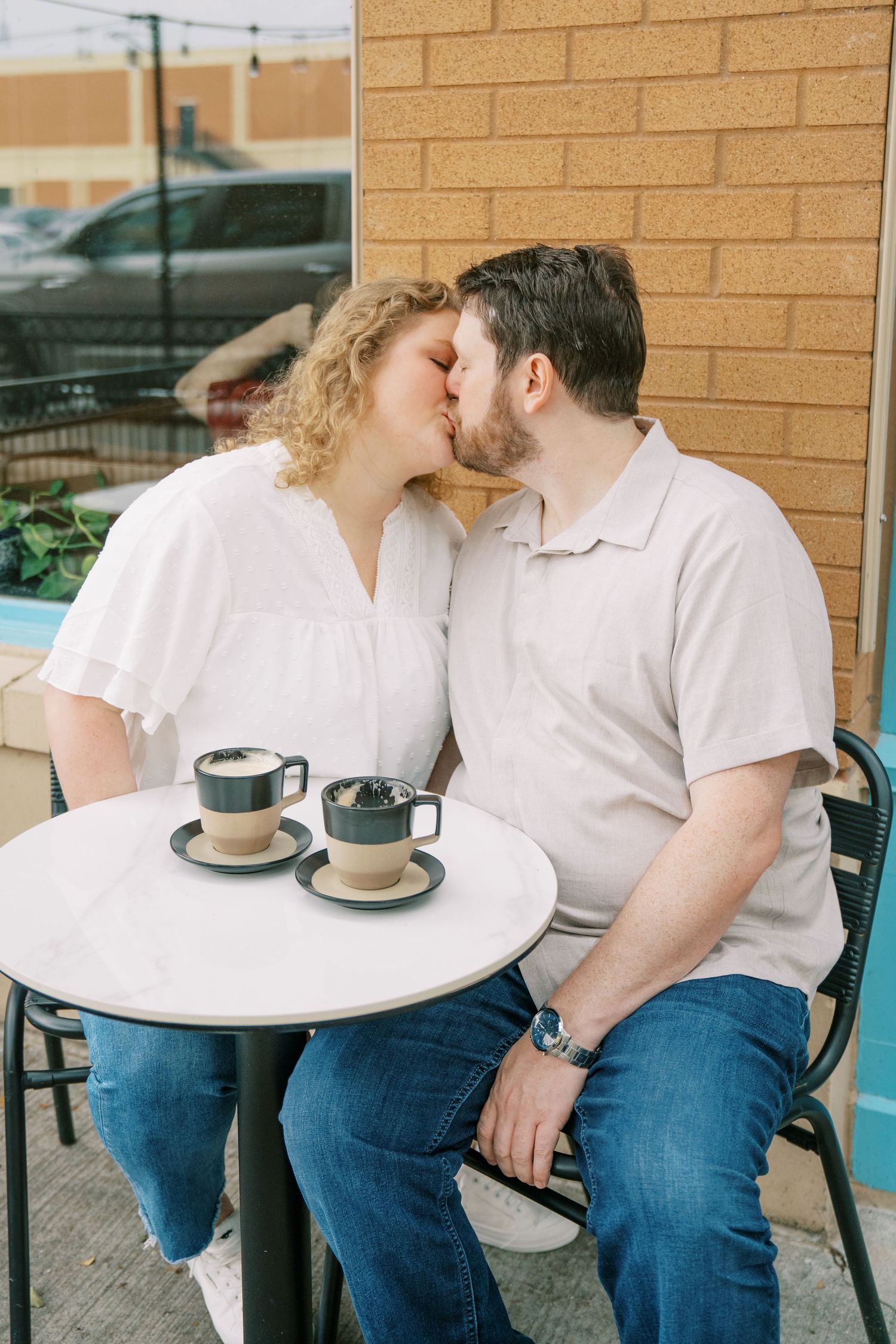Couple sharing a kiss at outdoor cafe table with two coffee cups between them.
