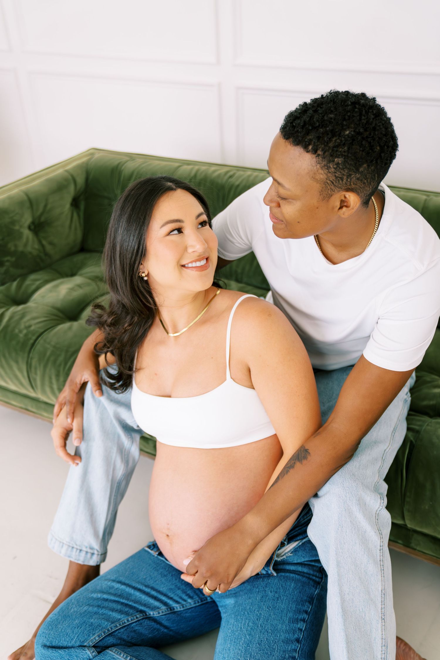 Happy pregnant woman and partner sitting by green velvet sofa, sharing a tender moment during maternity photo session.
