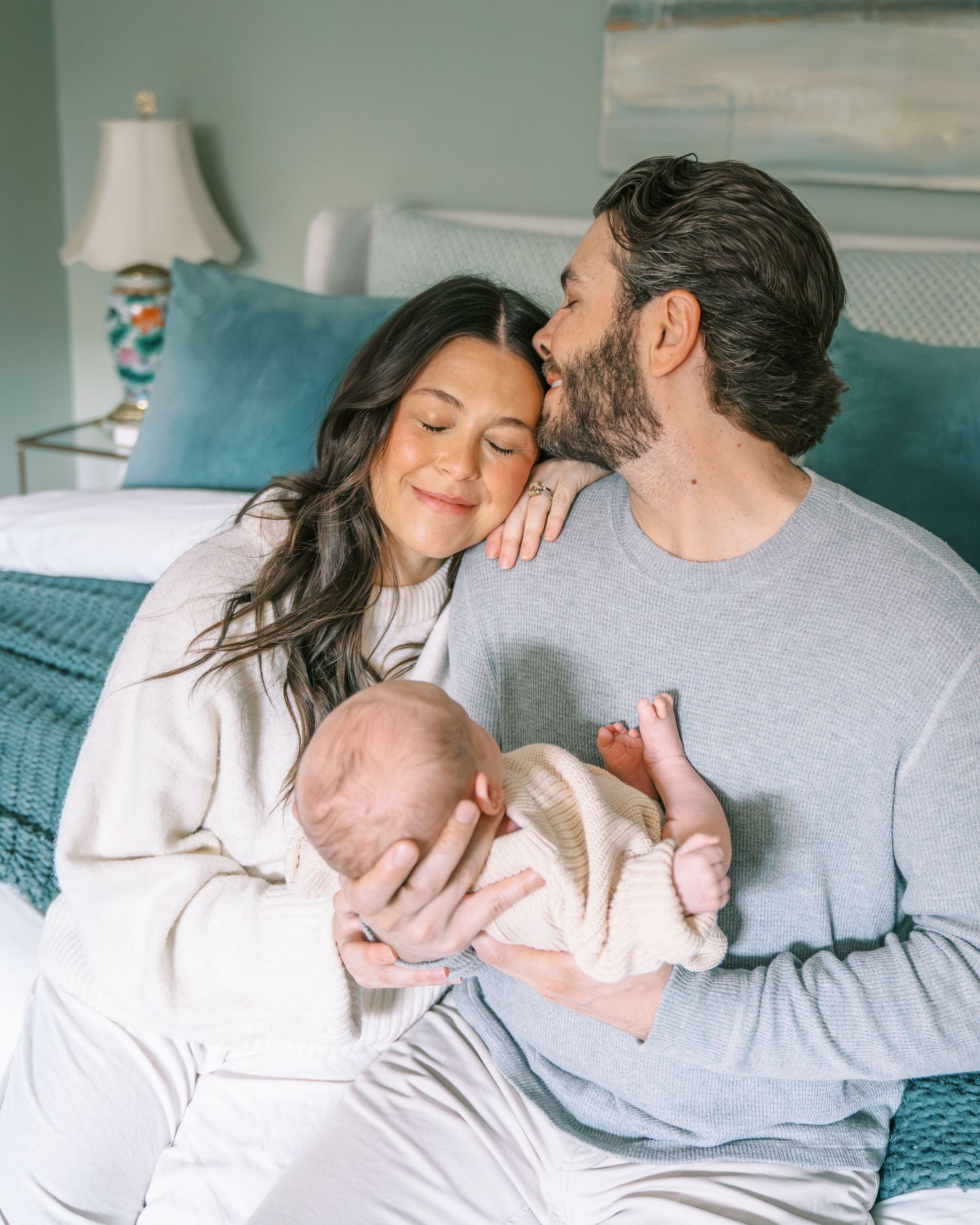A couple sits on a bed, smiling as they lovingly hold their newborn baby, with the man kissing the woman's head.
