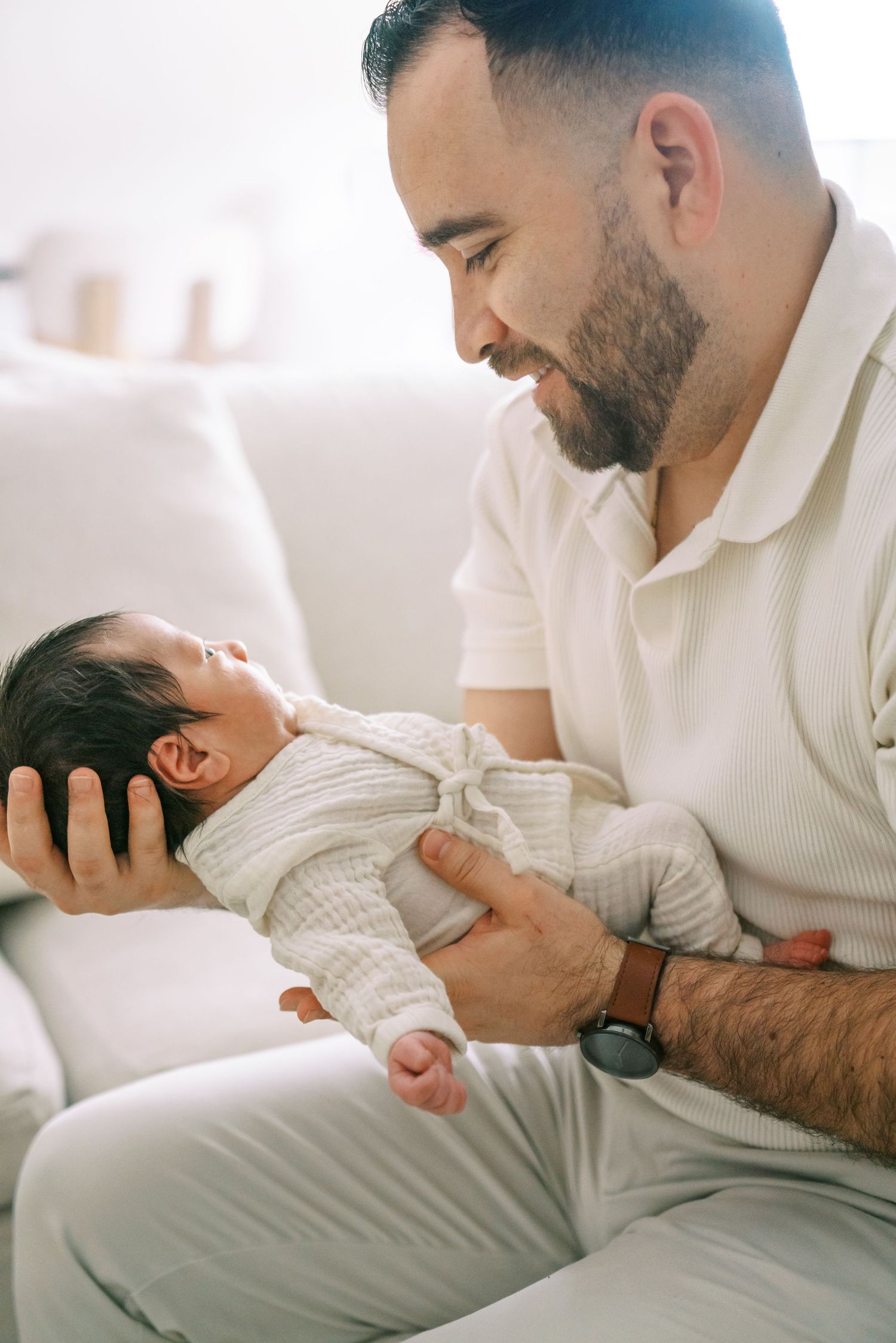 Bearded father tenderly holds his newborn baby in white outfit while sitting on a couch, gazing lovingly.