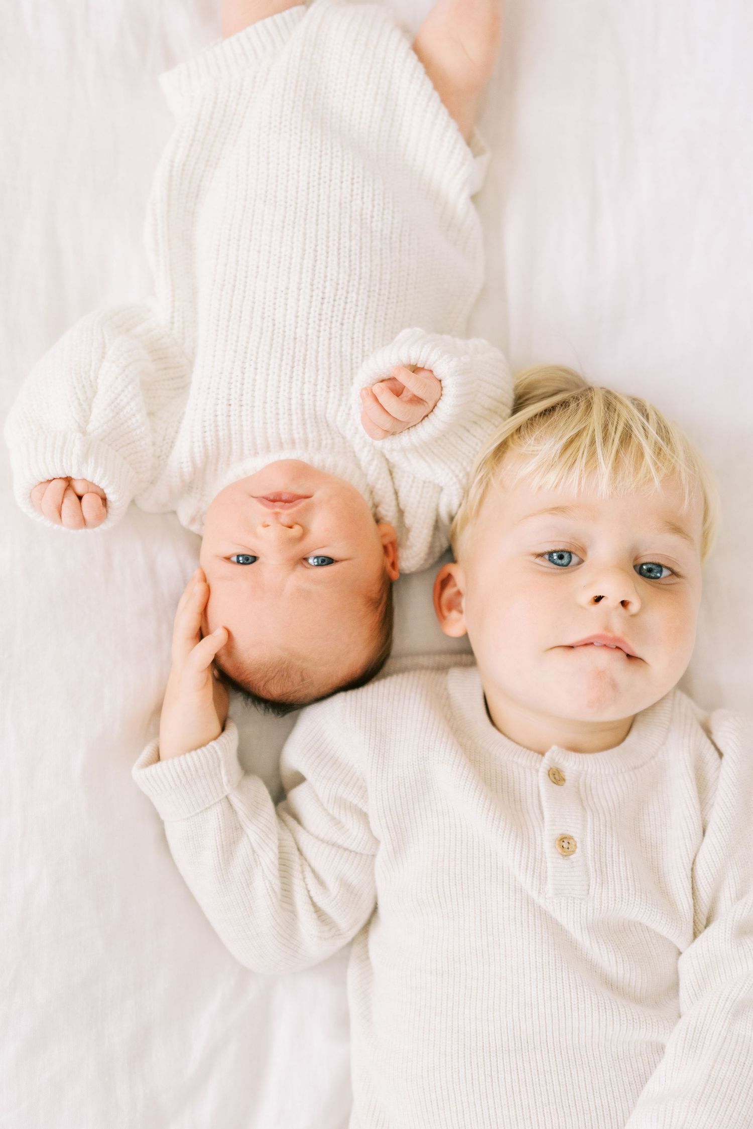 Two young children in white knit clothing lying together on white bedding.