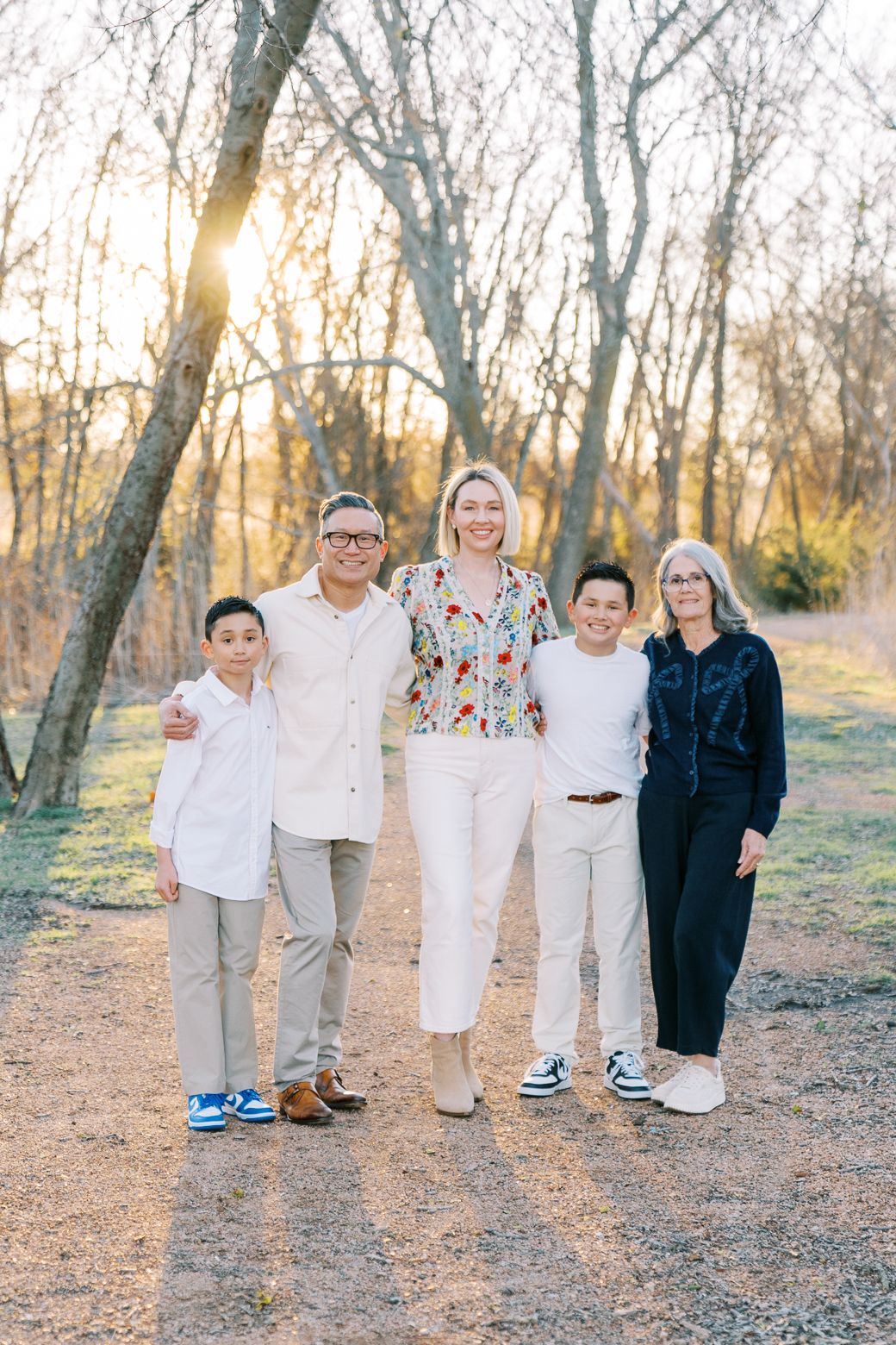 A group of people in coordinated white and neutral outfits pose together on a wooded path at sunset.