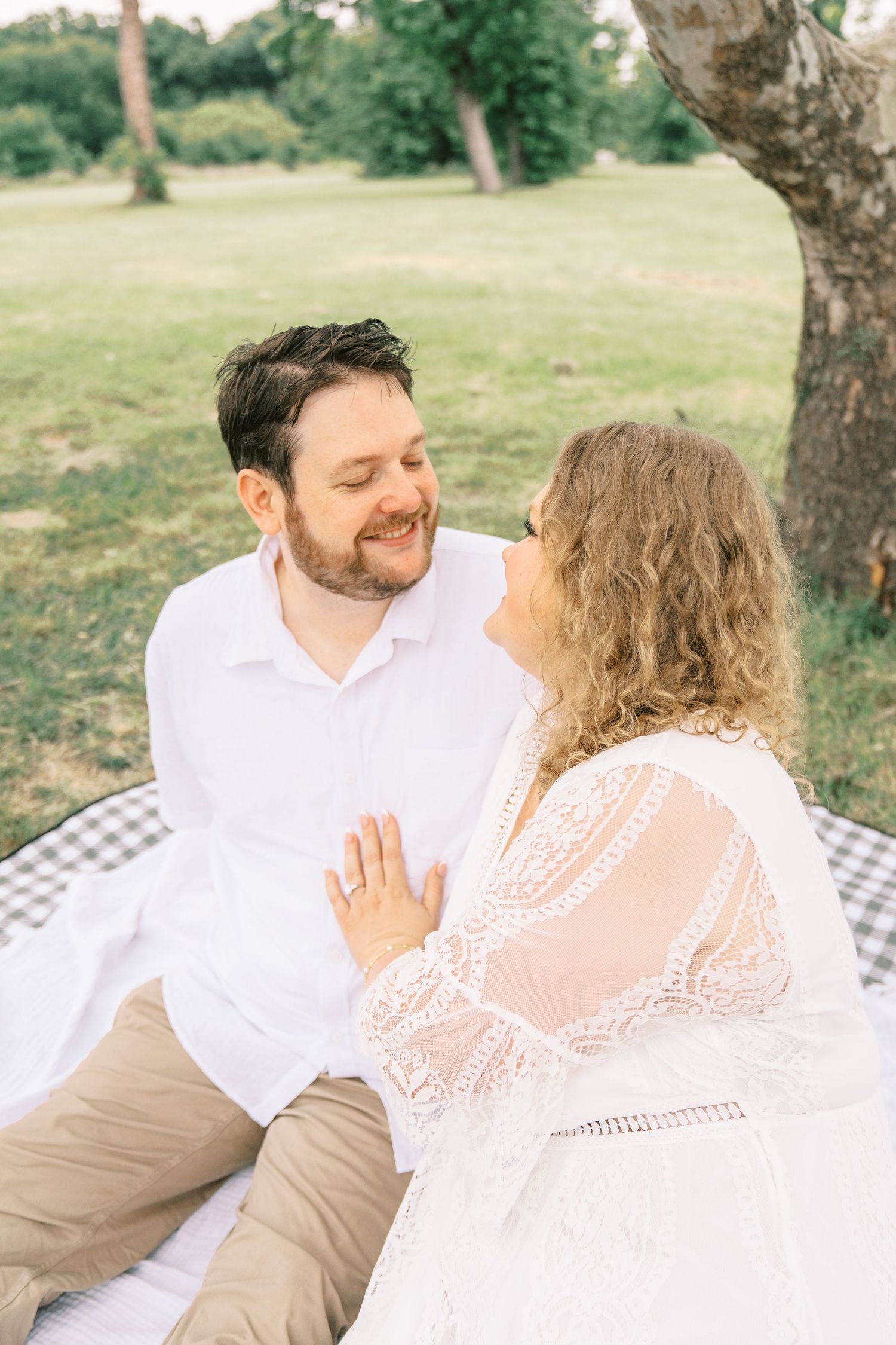 Smiling couple sitting on a blanket in a park, man in white shirt and woman in lace dress gazing at each other.