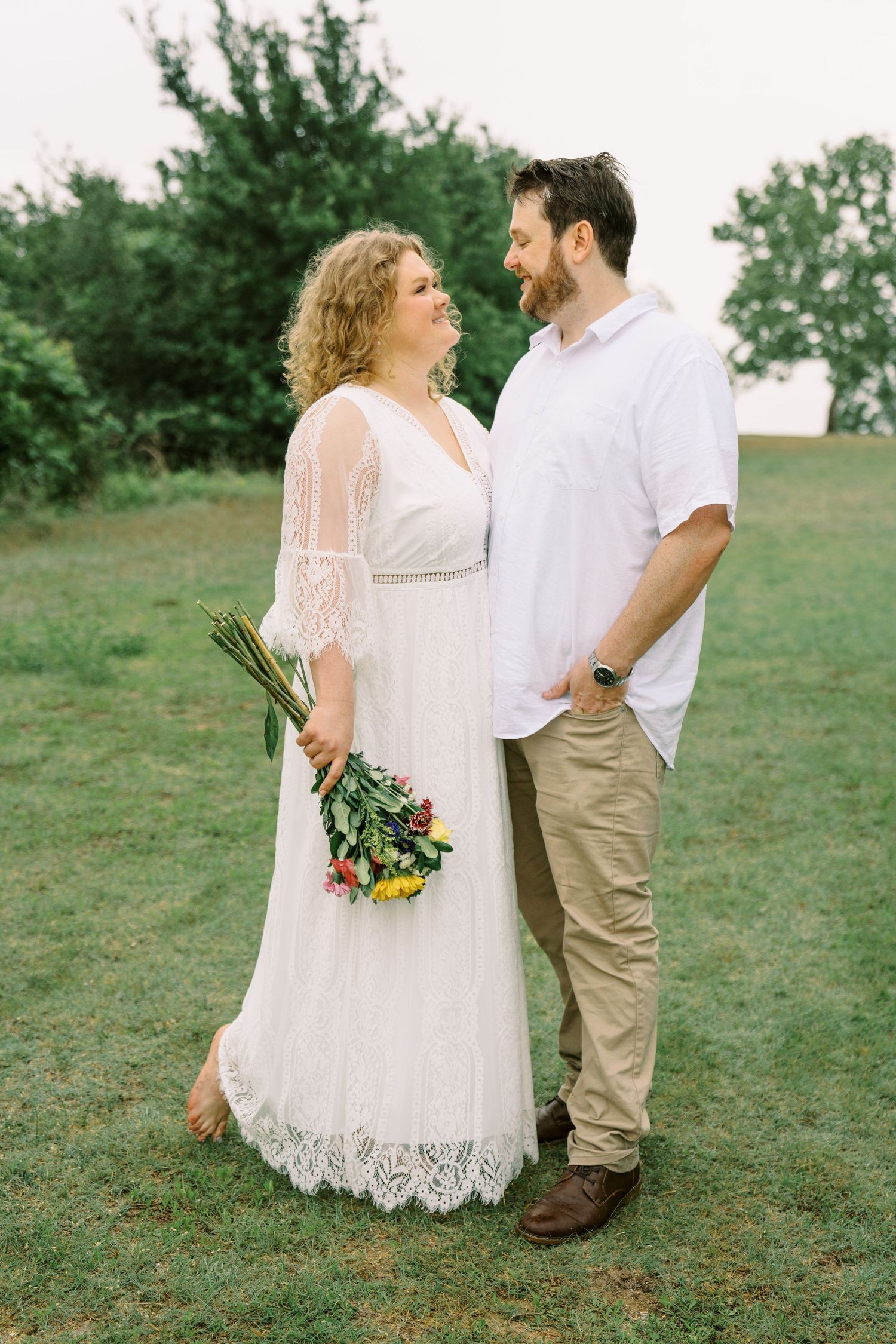 Smiling couple standing on green grass, woman in white lace dress holding colorful wildflower bouquet.