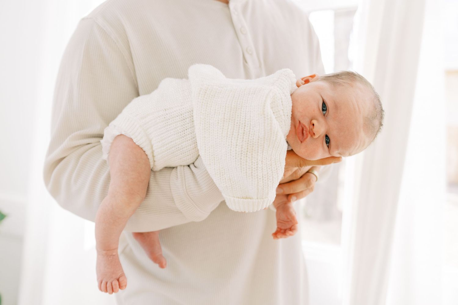 Adult cradling a newborn baby dressed in white knit outfit against a bright, airy white background.