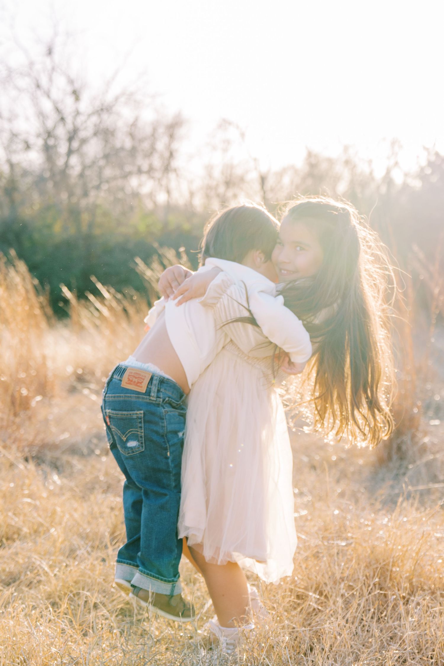 A mother and young child share a warm embrace in a sunlit golden field, backlit by soft natural light.