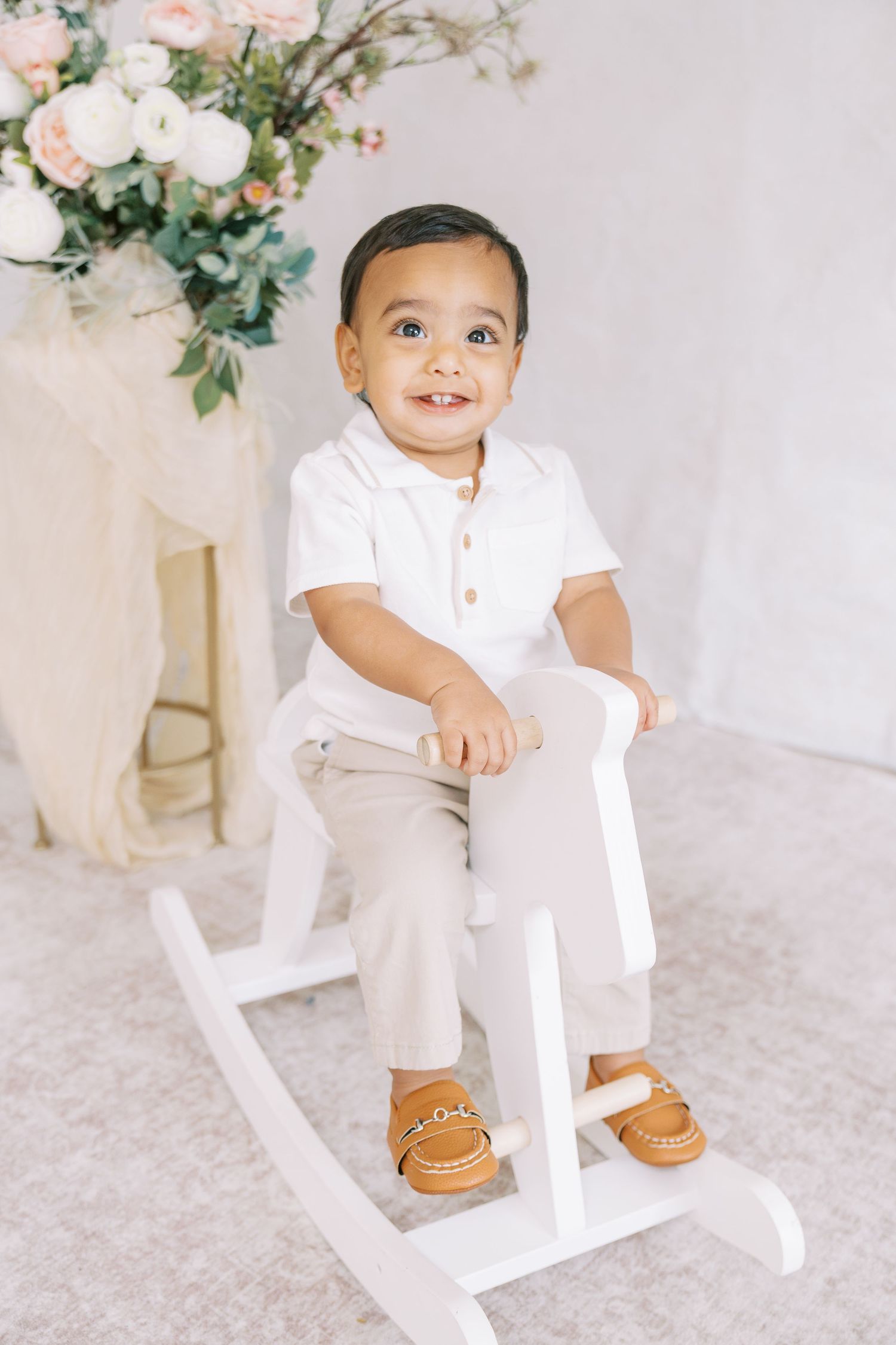 Smiling toddler boy in white polo and khaki pants sitting on a white rocking horse with floral decor behind him.