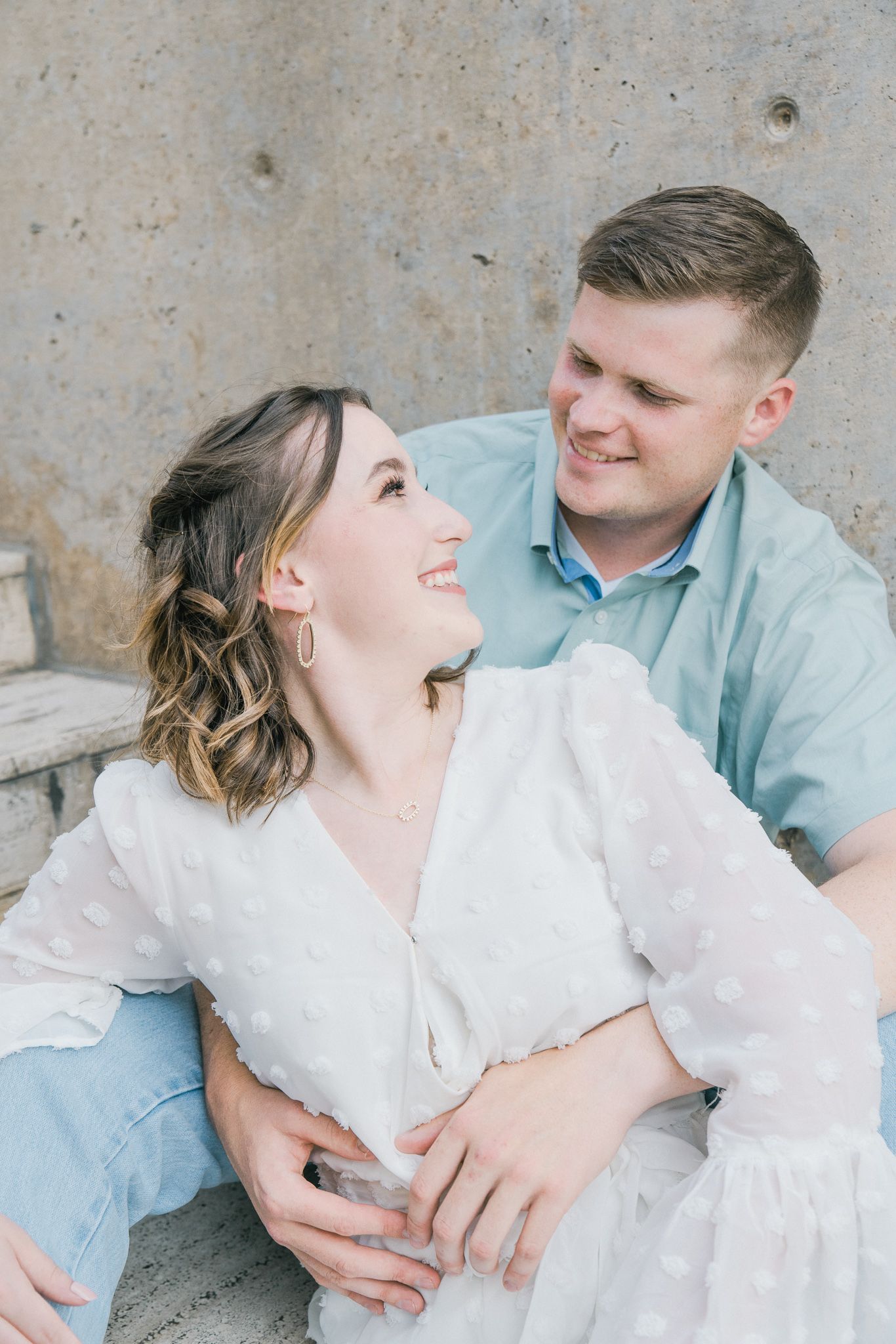 A romantic couple in light blue and white outfits share tender moments against a textured concrete wall.