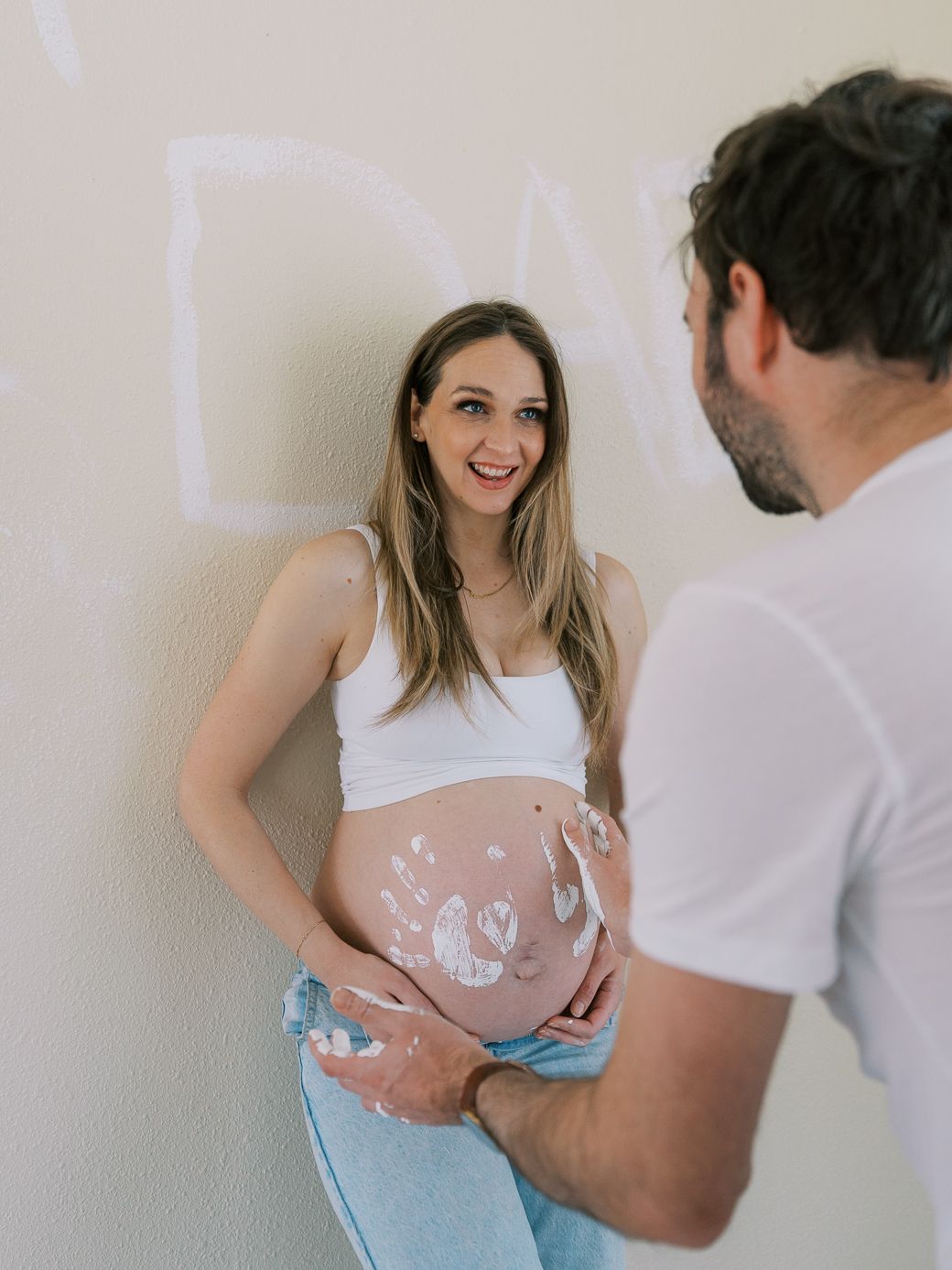 Couple creating heart shapes with hands on pregnant belly in casual white and denim outfits.