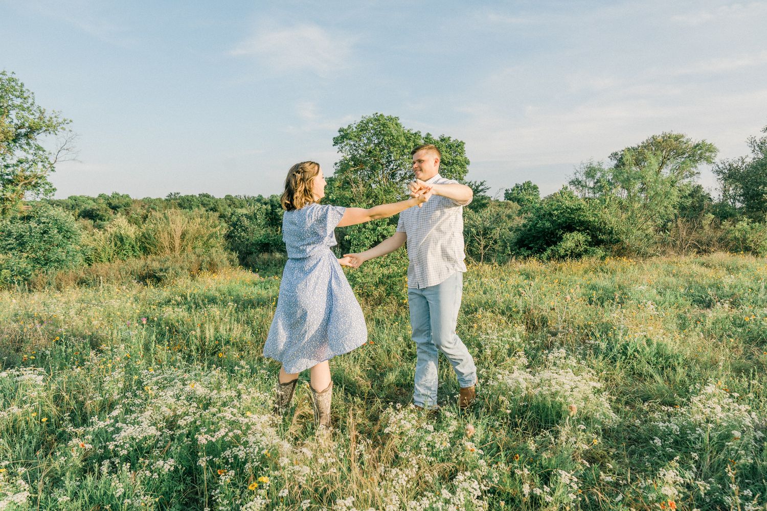 Two people in light colored clothing dance together in a sunlit meadow filled with wildflowers.