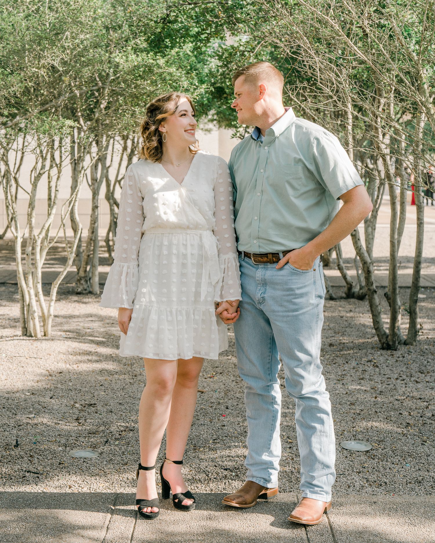A couple shares a joyful moment together on a tree-lined path, with her in a white dress and him in casual attire.
