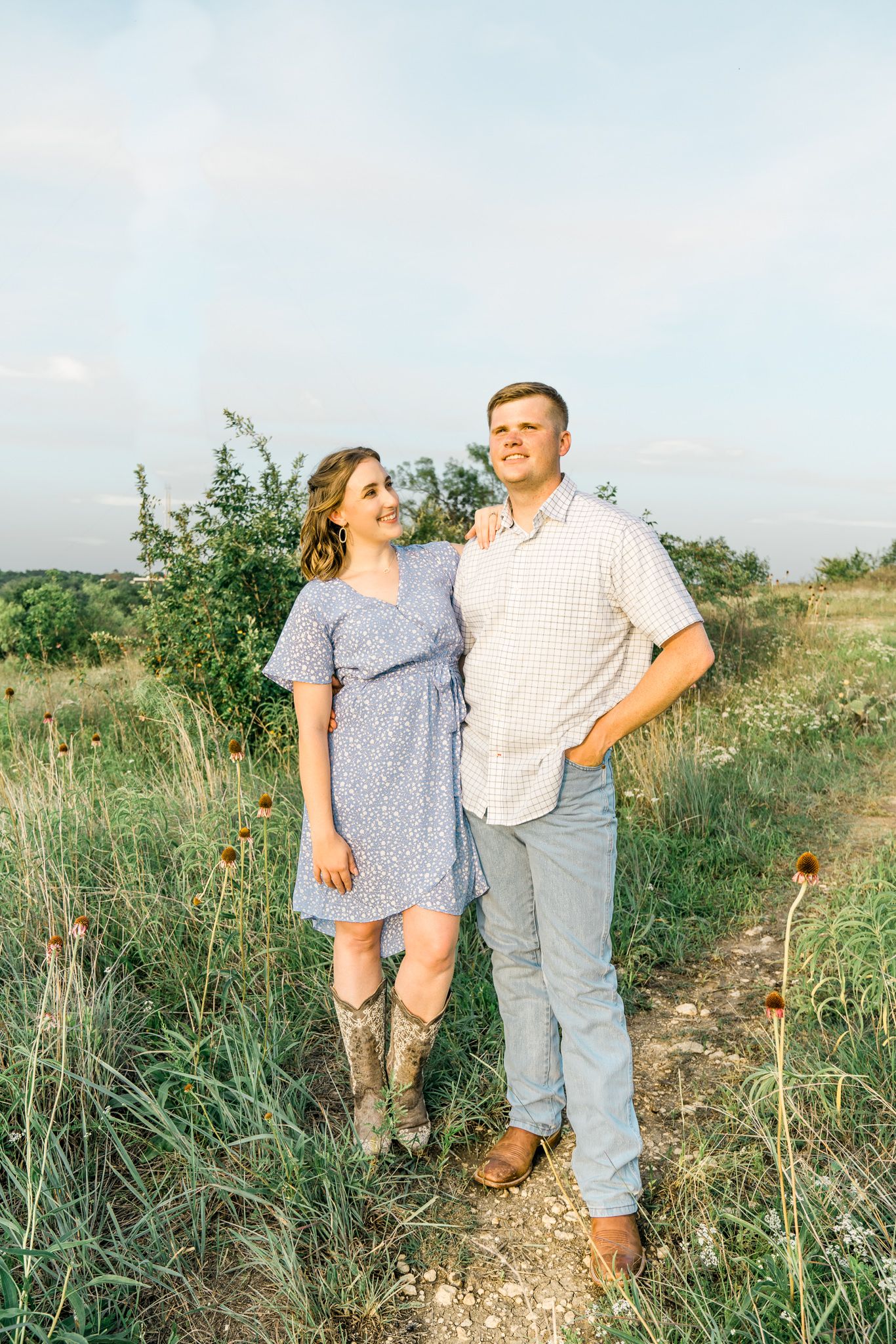 A couple stands together in a grassy field at sunset, wearing casual attire with cowboy boots and summer dress.