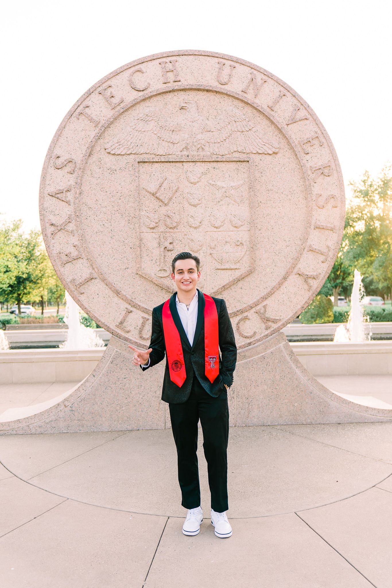 Graduate in black suit and red stole poses in front of architectural stone wall at university campus.