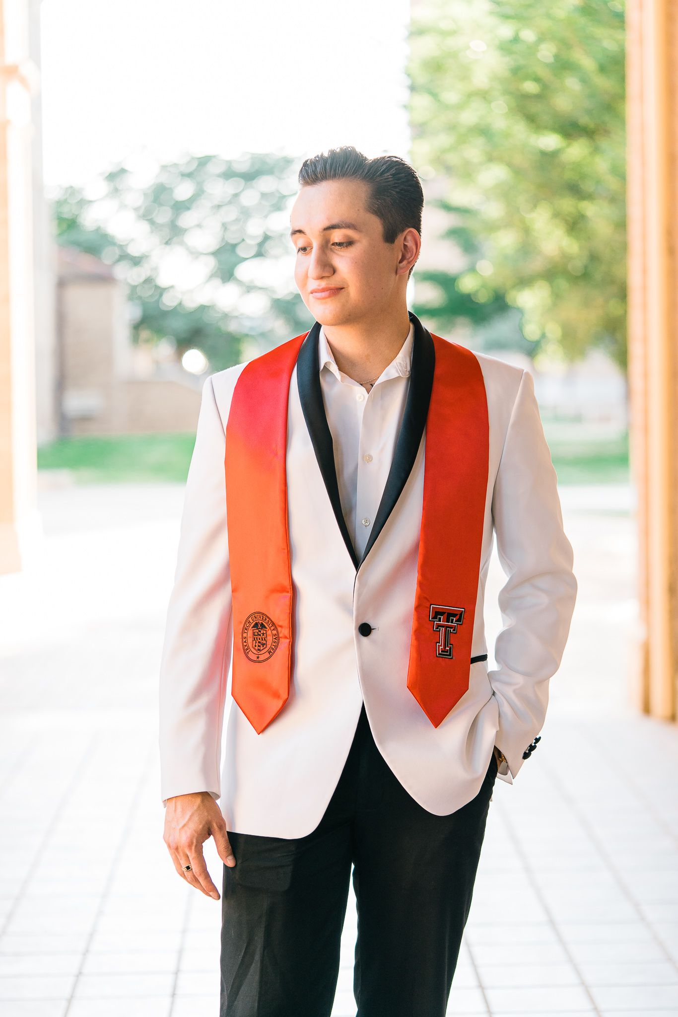 Graduate wearing white tuxedo jacket and red graduation stole smiles while walking down covered walkway.