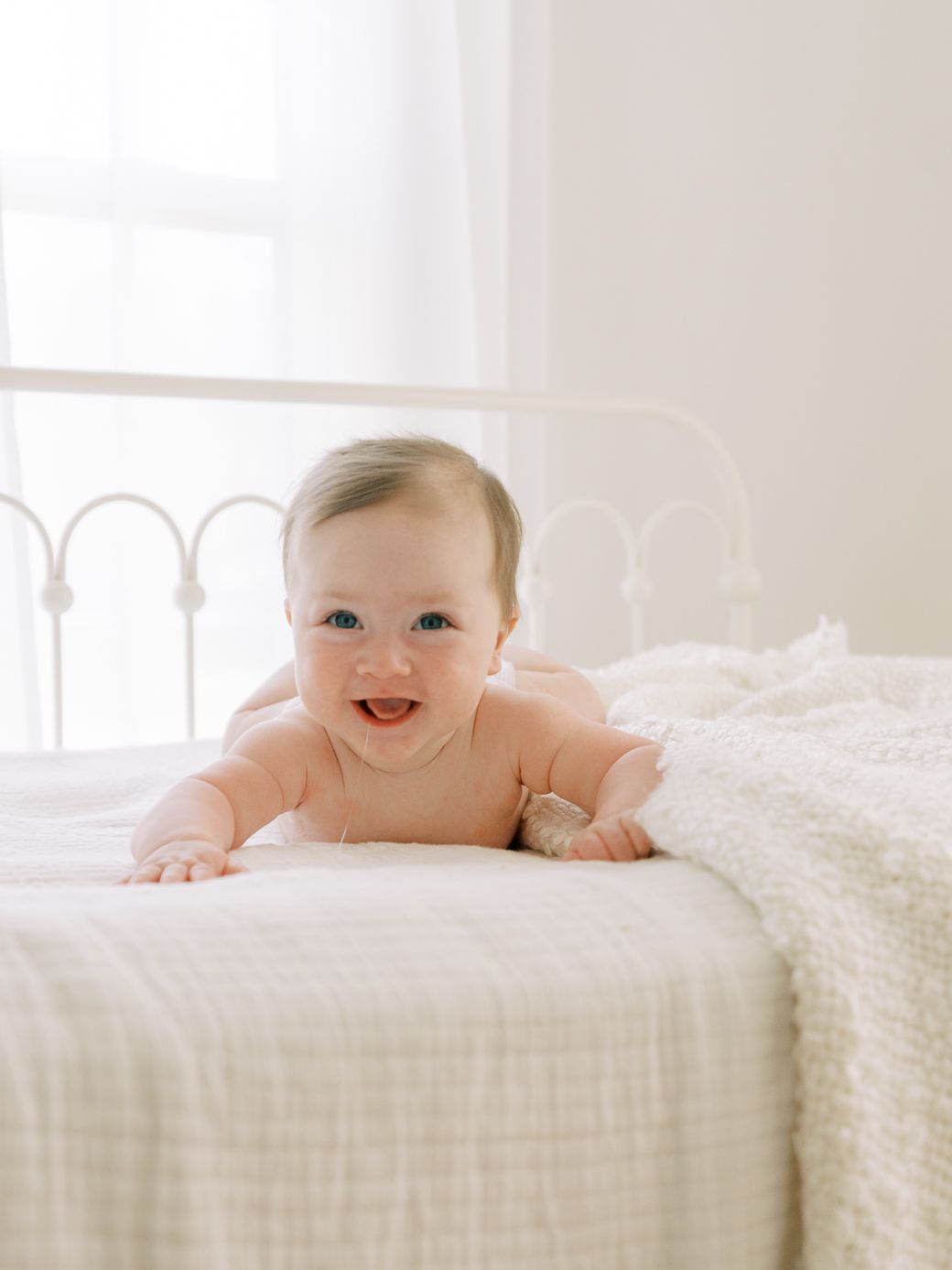 A happy baby lies on a white bed with soft textured bedding in a bright, airy room.
