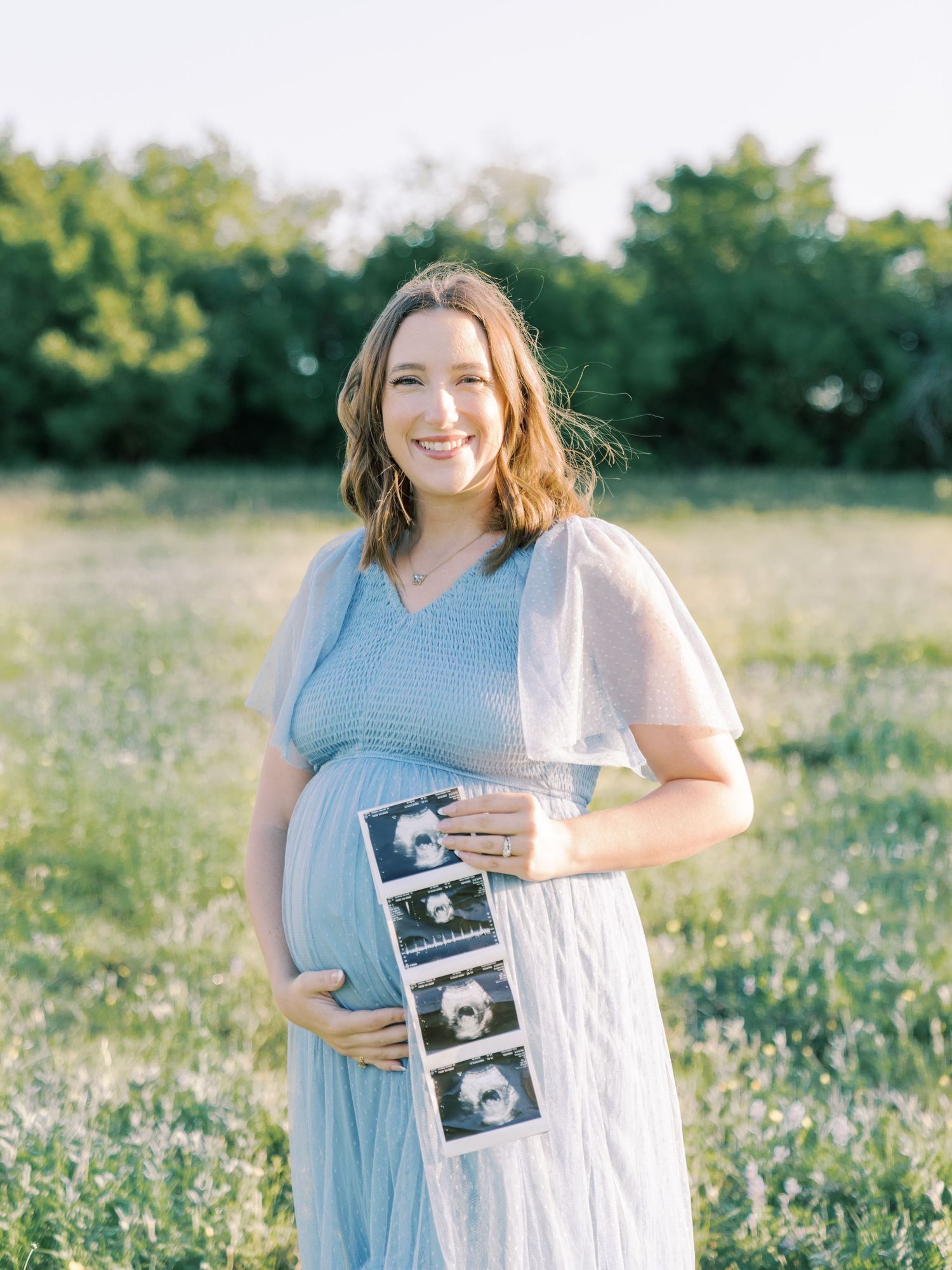 A woman in a light blue dress holds ultrasound photos while standing in a sunny wildflower field.