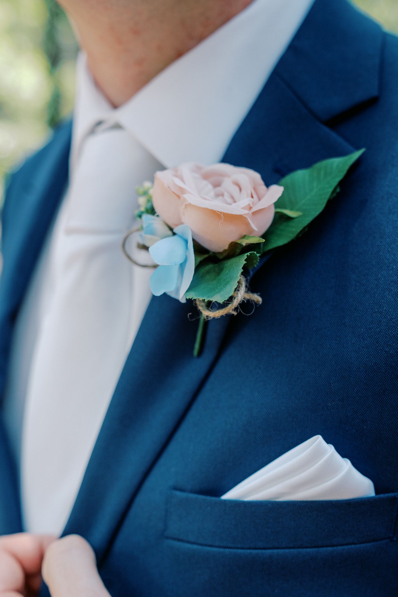 A light pink rose boutonniere with green leaves pinned to a navy blue suit jacket lapel.