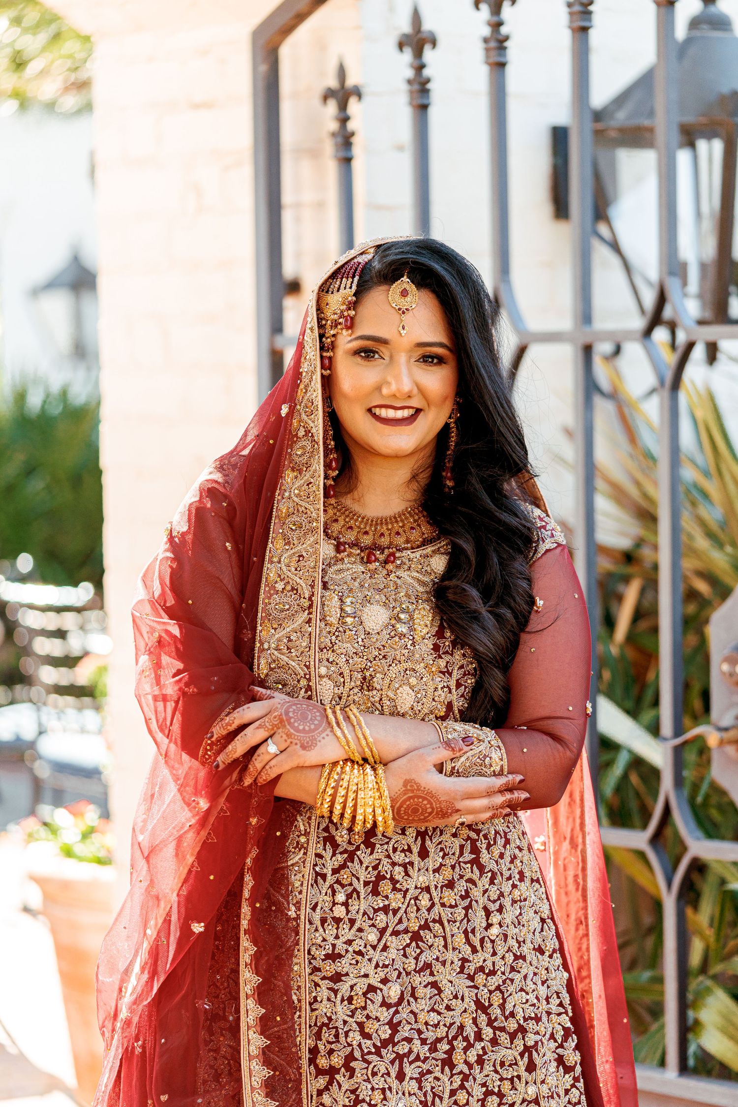 Bride wearing heavily embellished gold and red wedding attire smiles during outdoor portrait session.