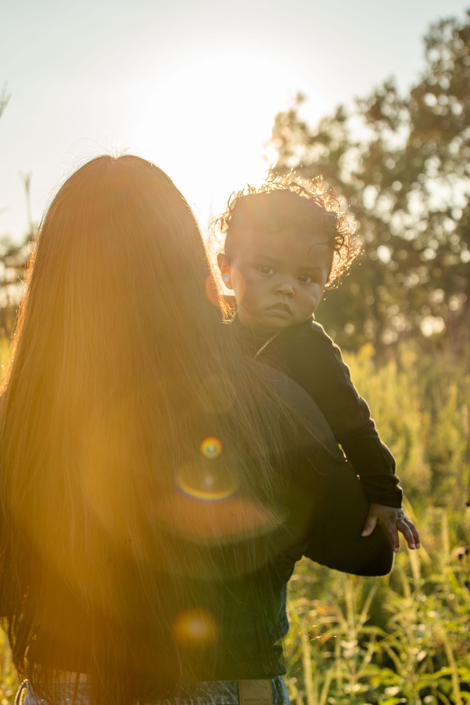 Silhouette figure in a black sweater stands in an outdoor field against a warm sunset with dramatic lens flare.