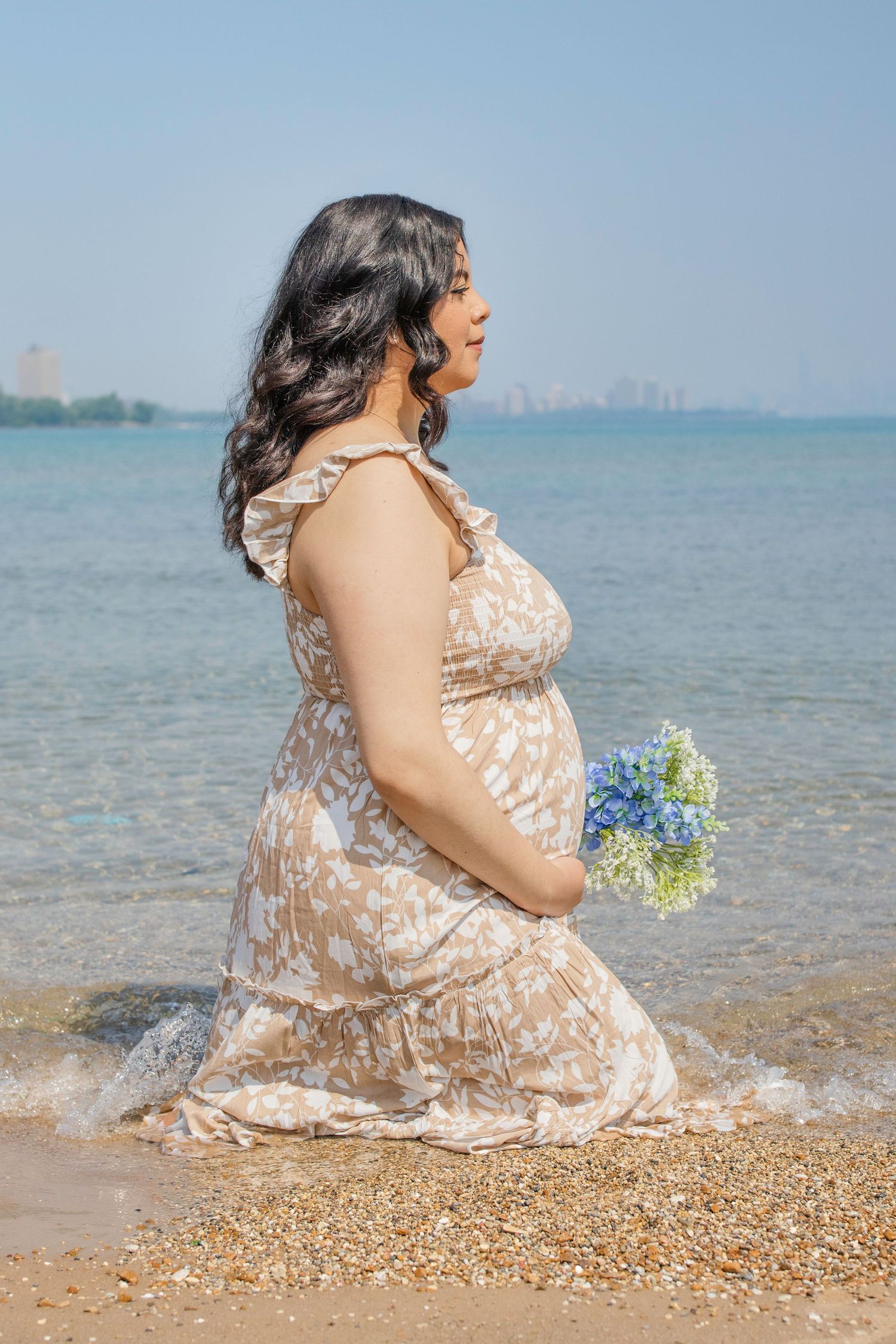 An expectant figure in a beige lace dress sits at the beach holding blue flowers with a city skyline in the background.