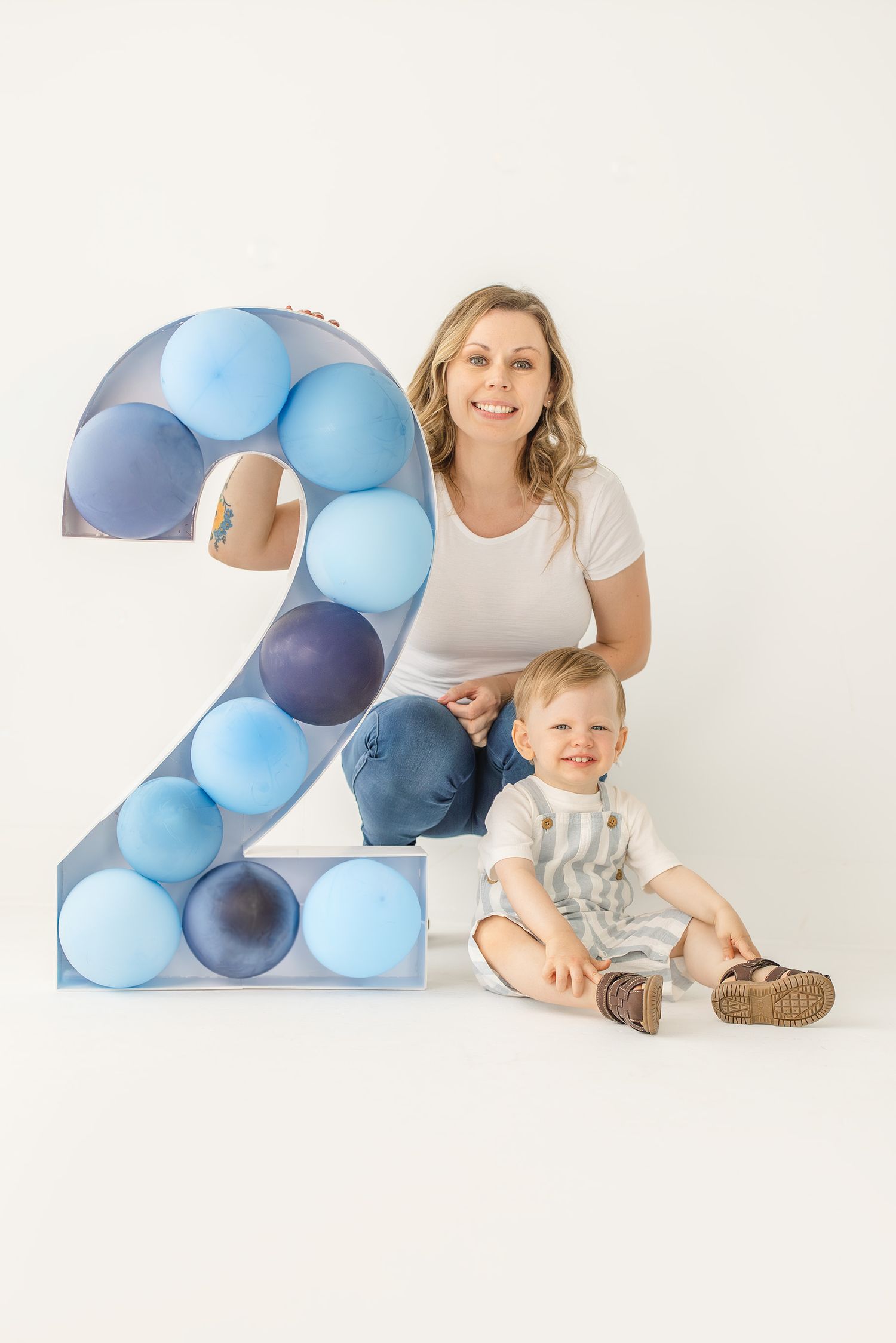 A smiling mother and toddler boy pose beside a large number 2 filled with blue balloons for a second birthday celebration.