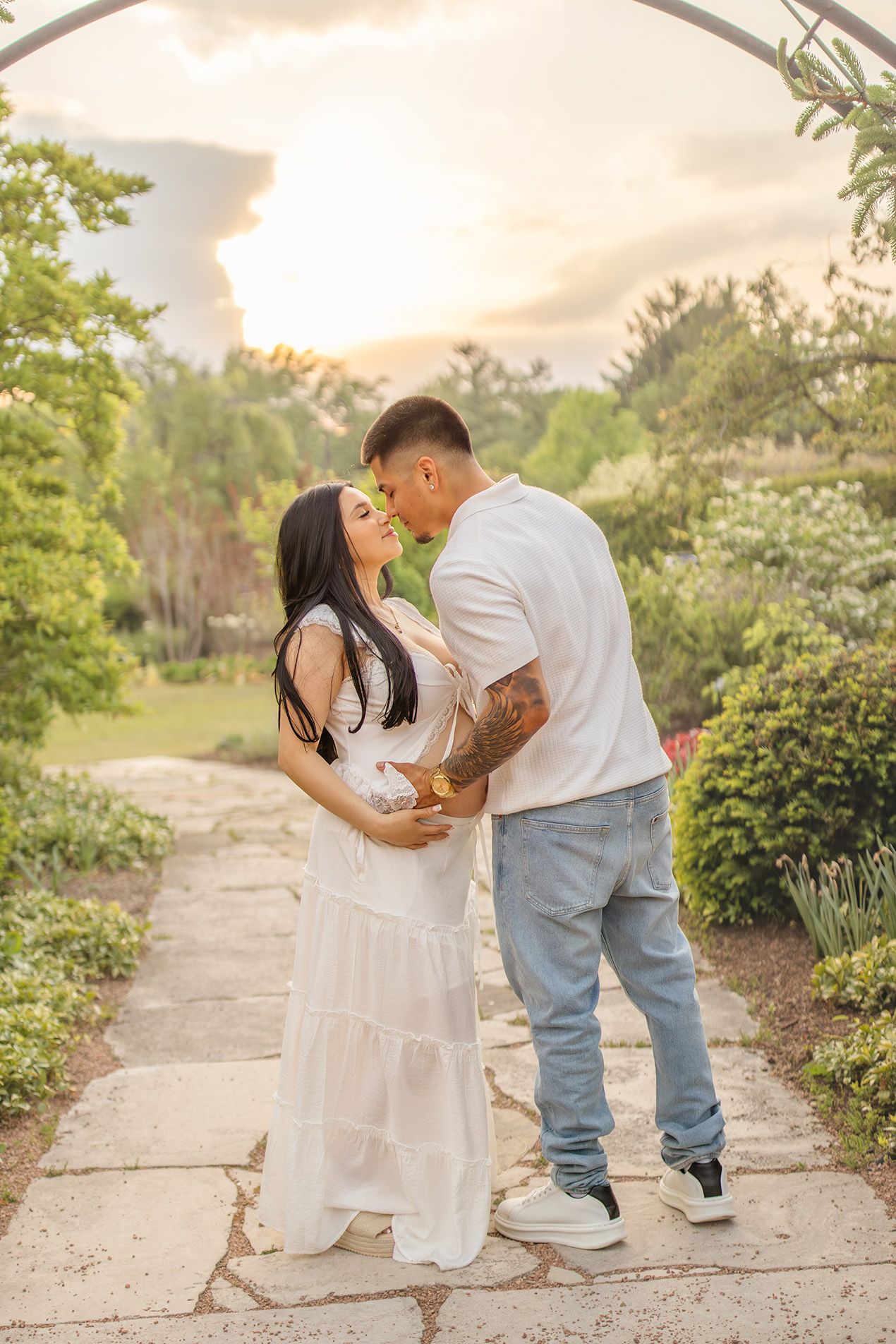 Couple sharing a romantic moment under garden arch at sunset, woman in white maternity dress with partner in white tee.