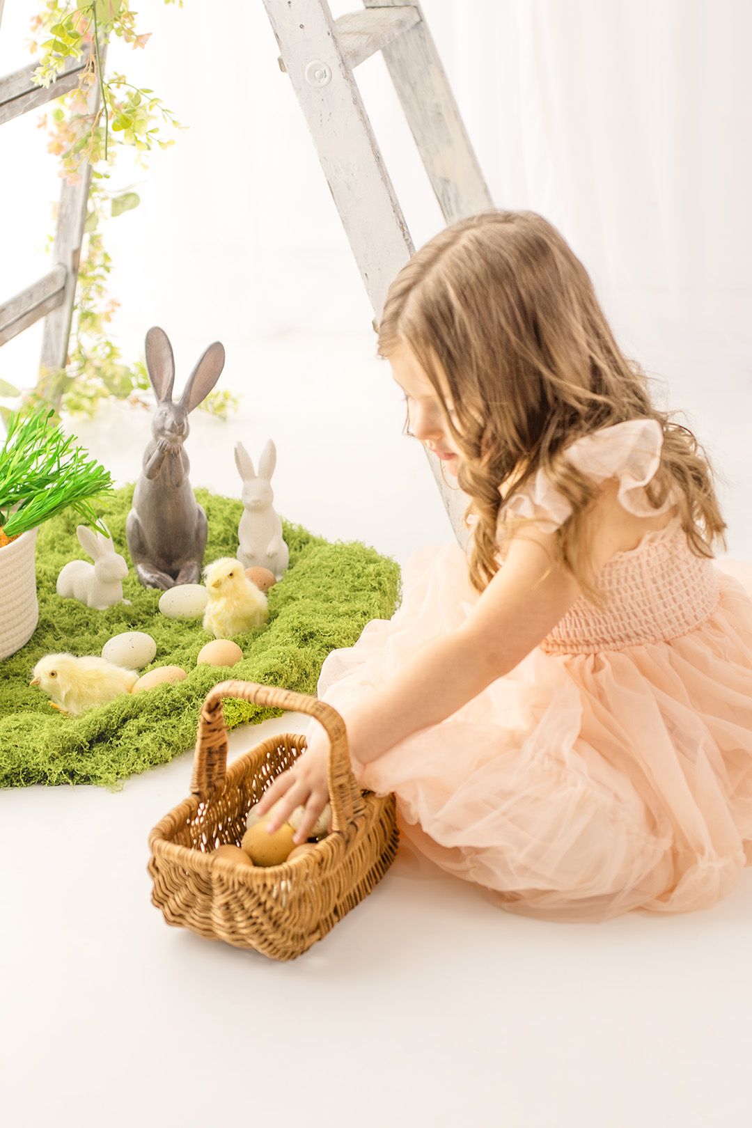 A child in a pink dress arranges Easter decorations with bunnies, chicks and a woven basket in a bright spring setting.