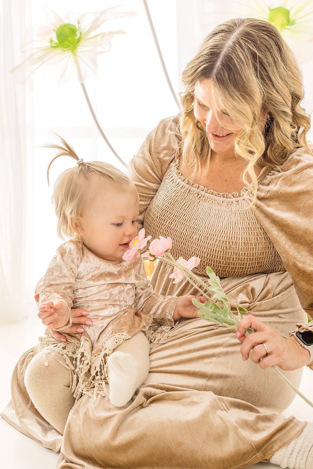Mother and child in matching beige outfits share tender moments while sitting together on white backdrop.