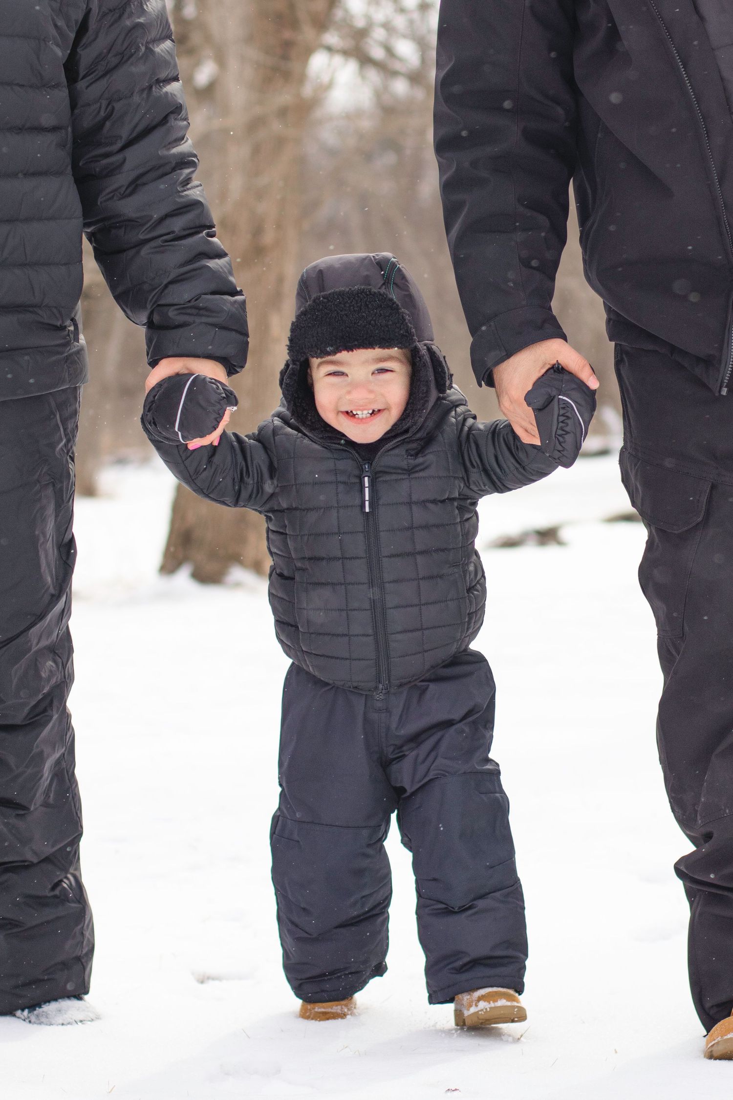 A small child in a black winter snowsuit smiles while holding hands in the snow with adults.