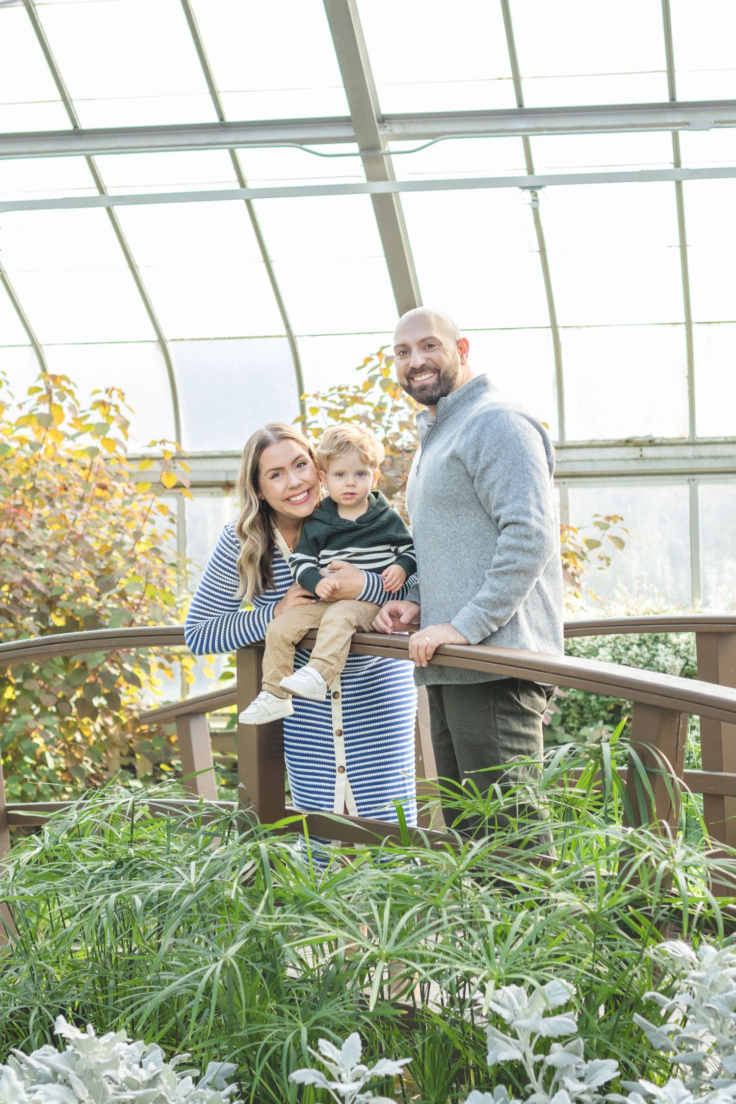 A family poses together on a wooden walkway inside a sunny greenhouse.