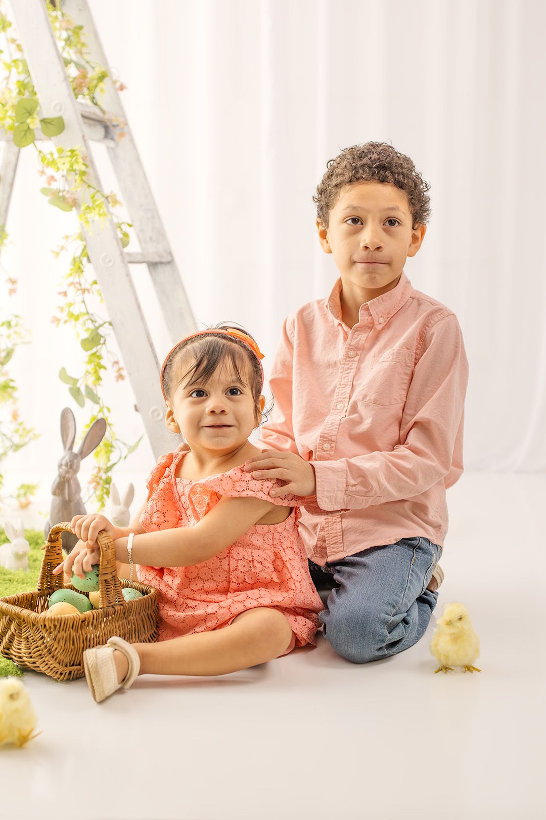Two children in pink outfits pose with Easter basket, eggs, bunny, and chicks in a bright studio setting.