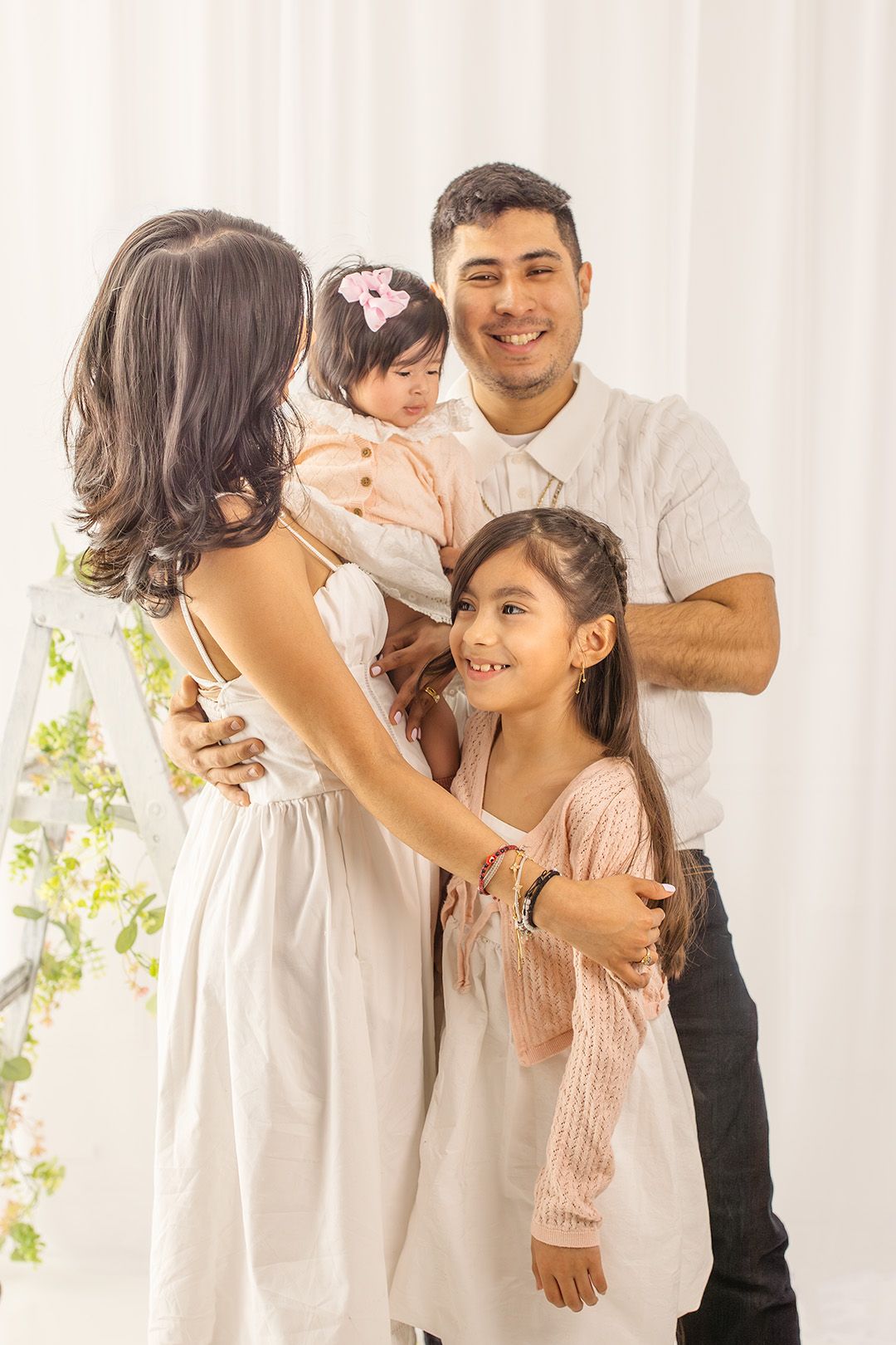Happy Hispanic family of four posing together in a bright white studio, parents holding two young daughters in matching light outfits.