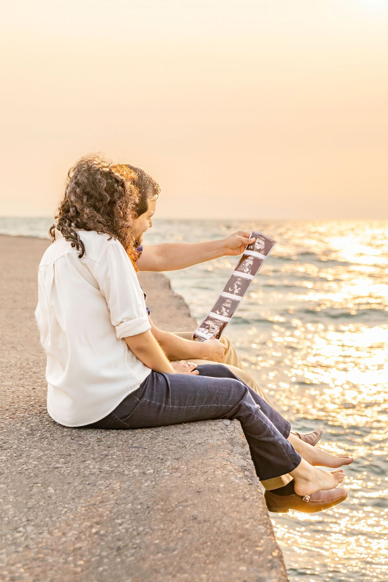 Person in white shirt and dark pants sits by beach at sunset while looking at ultrasound photos.