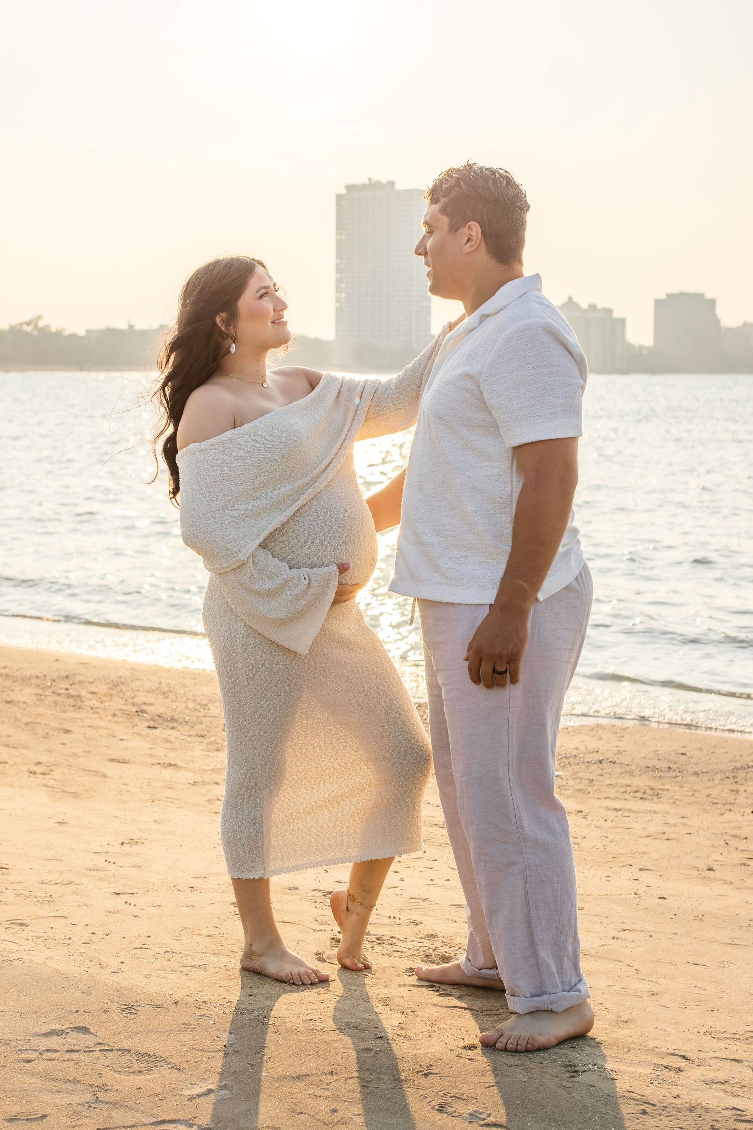 Two people in white outfits share romantic moments on beach at sunset with buildings in background.