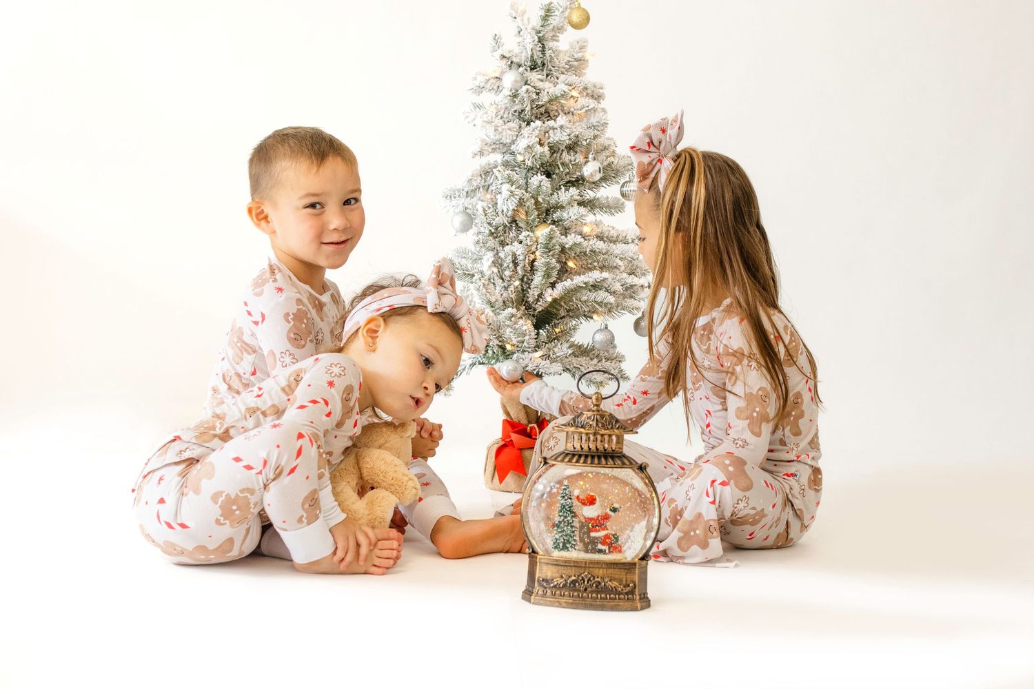 Children in matching holiday pajamas sit together near decorated Christmas trees and festive decor in a bright studio setting.