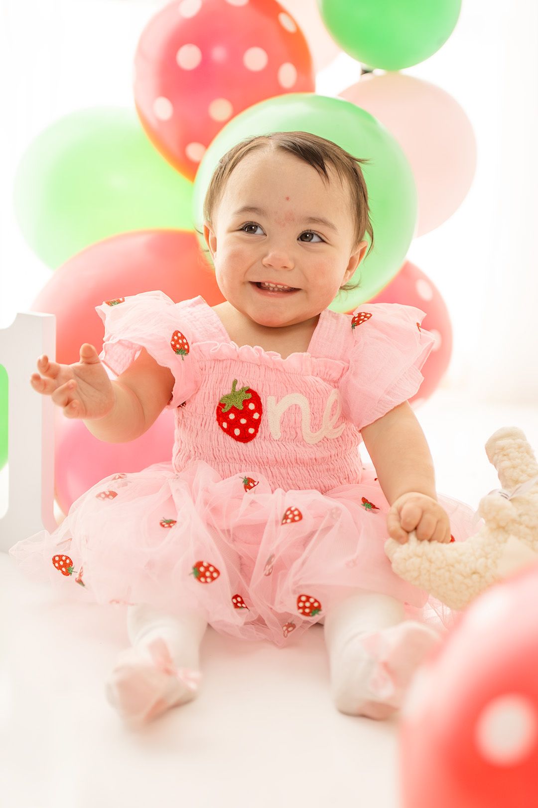 A smiling baby girl in a pink strawberry dress celebrates her first birthday surrounded by colorful balloons.