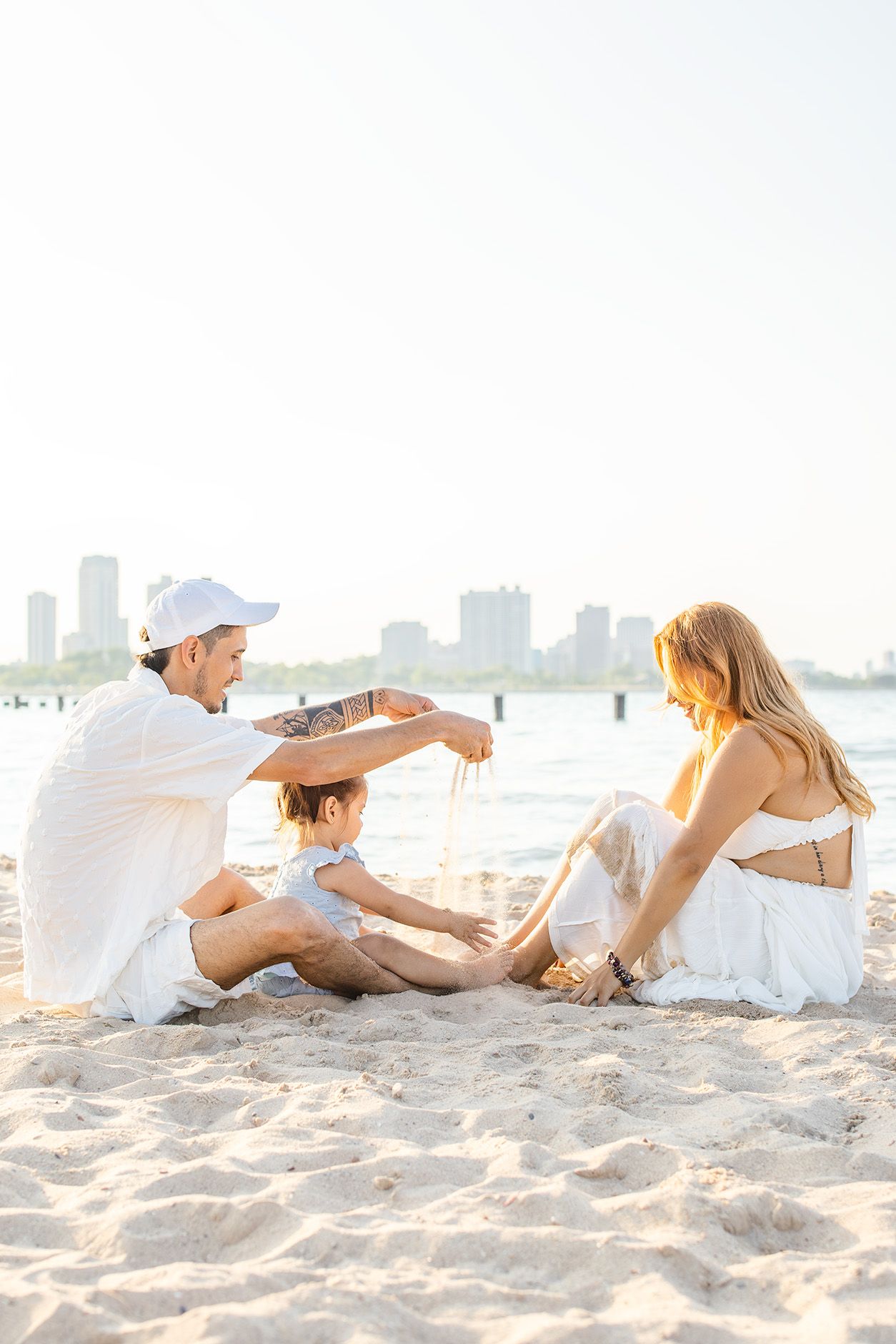 A happy family sits on a sandy beach, with a father pouring sand over his child's head.