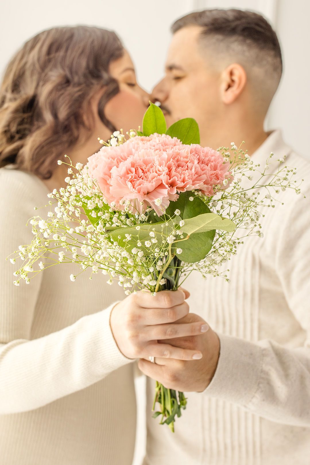 Couple kissing behind a bouquet of pink carnations and white baby's breath flowers.